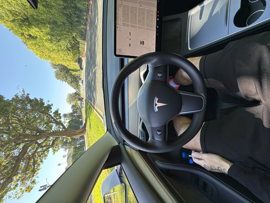 From inside a Tesla, a view of a tree-lined street on a sunny day. The dashboard screen displays a map. The steering wheel is visible. A person's legs are visible, clad in black shorts, along with an arm that has a tattoo, reaching toward the car's controls. The car appears to be moving slowly.