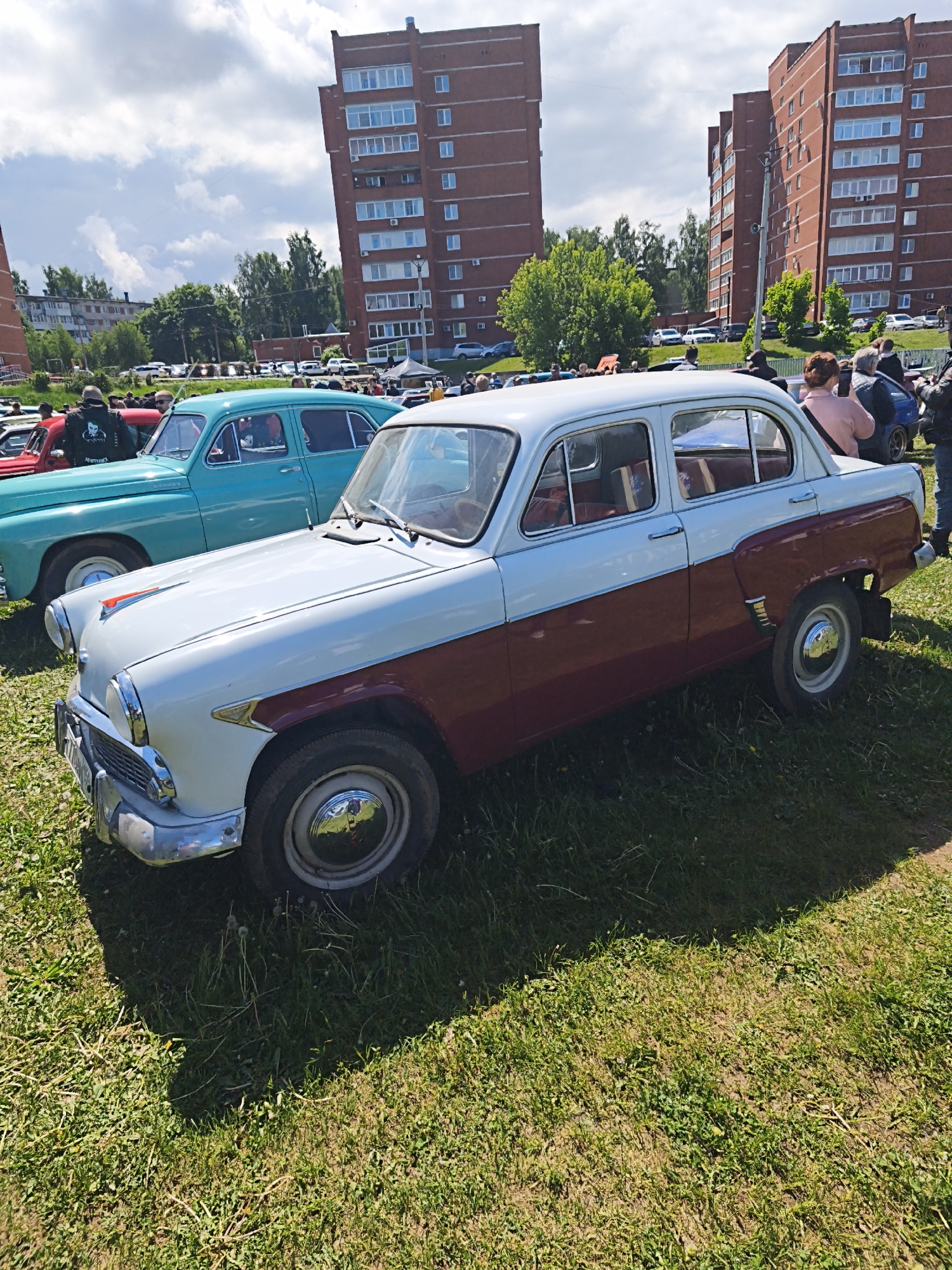 The image shows a two-tone white and maroon Moskvich 407 car parked on a grassy area in Chaikovsky, Russia. Another vintage teal car is parked behind it. People are gathered around the cars, some appearing to be taking pictures. The backdrop includes multiple-story brick apartment buildings, trees, and other vehicles parked in the distance, indicating an outdoor event or car show setting. The sky is partly cloudy.