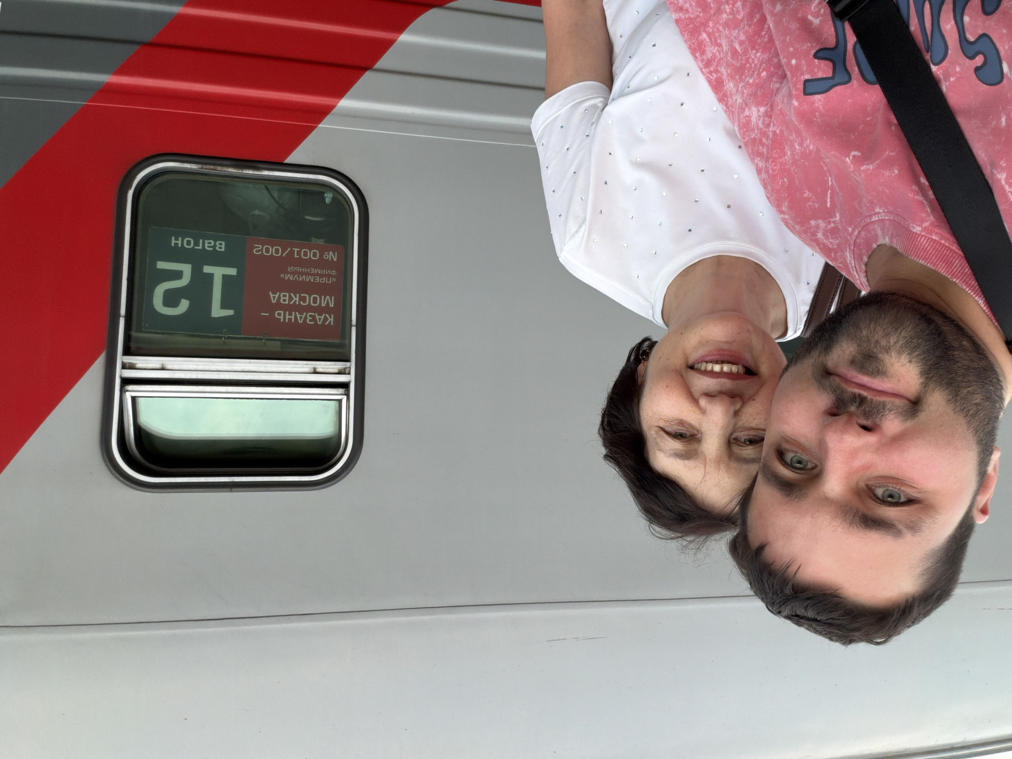 A selfie photograph features two people in front of a train. The person on the left has short dark hair and is wearing a white t-shirt. The person on the right has a beard and is wearing a pink sweatshirt with blue lettering. The train is light gray with a red stripe and the number 12 is visible in a window. Text in the window indicates the train is traveling from Kazan to Moscow. The photograph appears to be taken inside of the city of Moscow, Russia.