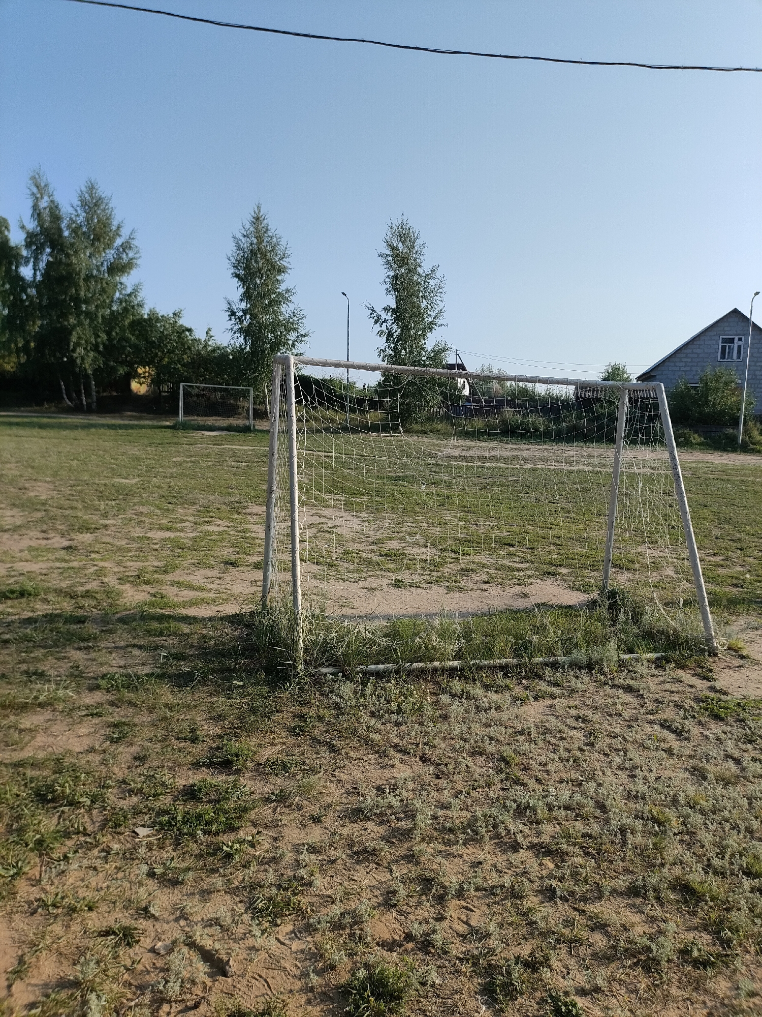 The image depicts an outdoor soccer field with two goals. The field appears somewhat worn, with patches of grass and exposed dirt. In the foreground, a larger soccer goal with a net is visible, positioned slightly off-center to the right. The goalposts are white, and the netting is white and appears to be in good condition. In the background, a smaller soccer goal with a net is visible further down the field. Trees line the perimeter of the field. Behind the trees, a gray, gabled house with a window is visible, suggesting a residential area. A streetlight is also present near the house. The sky is clear and blue, indicating fair weather. The overall scene suggests a simple, perhaps rural, sports facility within the city of Petropavl, Kazakhstan, likely a public or community area. There are no people or animals visible in the image.
