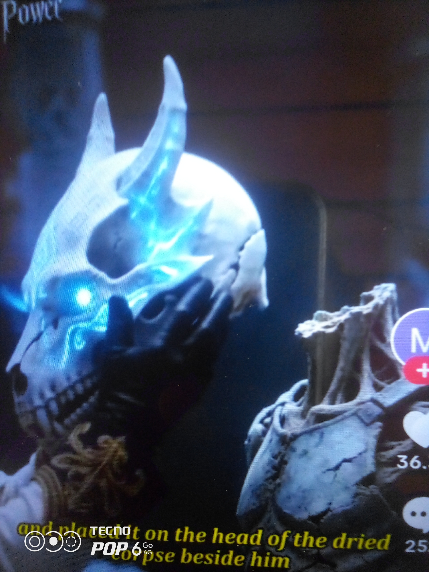 The image shows a close-up of a skull-like mask with glowing blue eyes and horns, with a portion of another skull-like object to the right. The background is dark and out of focus, with some text overlays visible. One overlay reads "Power" in the upper left corner. Another overlay at the bottom includes the text "and put it on the head of the dried" and "pose beside him", along with "TECNO POP 6 Go 4G". There are also social media icons and numbers visible on the right side of the image, suggesting it might be a screenshot from a video or social media platform. The overall scene is dark and somewhat eerie, with the glowing elements of the mask being the most prominent. The location in the city of Howayi, Nigeria, is not depicted or implied in the image.