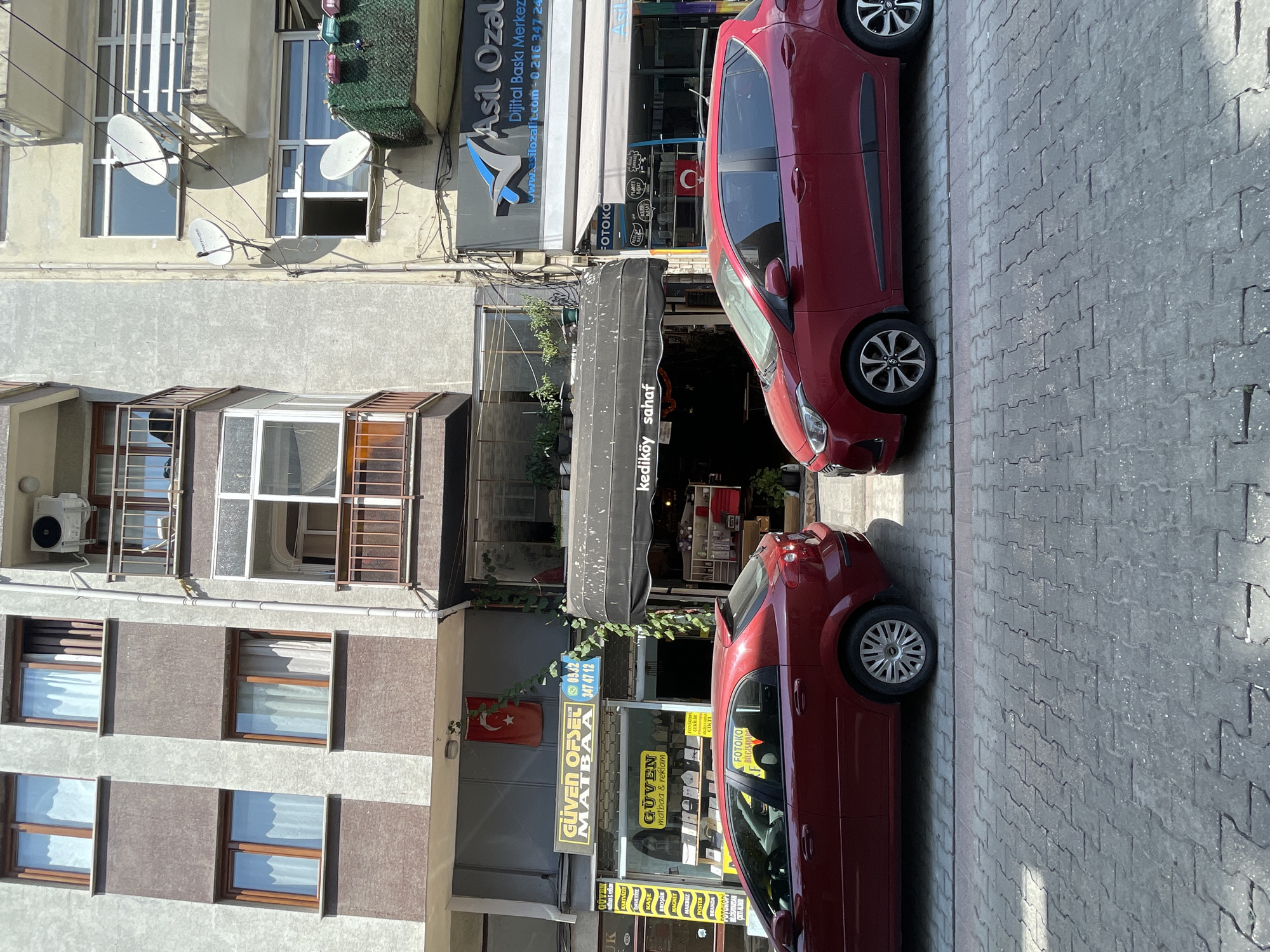 This is a street-level, eye-level, medium shot of a street scene in Kadıköy, Turkey. The buildings are multi-story, with a mix of residential and commercial spaces. Two red cars are parked on the street, one in the foreground on the left and another slightly further back on the right. Several businesses are visible on the ground floor, including a print shop with a yellow sign that reads "GÜVEN OFSET MATBAA" and a shop with a black awning that says "kediköy sahaf". There are satellite dishes mounted on the side of one building, and plants are visible on balconies. The street is paved with interlocking grey stones. The overall scene depicts a typical urban environment in a Turkish city.