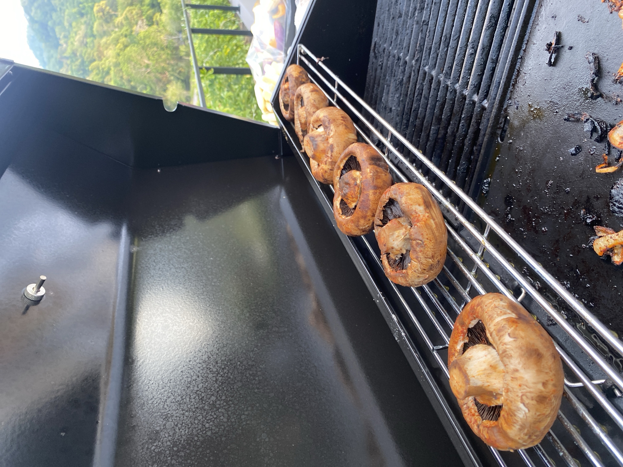An outdoor, brightly lit shot captures a close-up of mushrooms grilling on a black barbecue. Six large, brown mushrooms, likely Portobello, are neatly lined up on a chrome upper warming rack of the grill, showing various stages of browning from cooking. The grill's main surface is a mix of a standard grate and a flat griddle-like section, both appearing used with some charred food remnants and grease visible. The body of the grill is made of textured black metal, with a small silver pin or knob visible on its left side.
In the background, out of focus, lush green foliage and trees suggest a natural, outdoor setting, possibly hilly or mountainous, consistent with the tropical environment of Choeng Thale, Thailand. The bright, even lighting indicates it is daytime with clear weather. No people or visible text are present in the image. The scene focuses entirely on the cooking process of the mushrooms on the grill.
