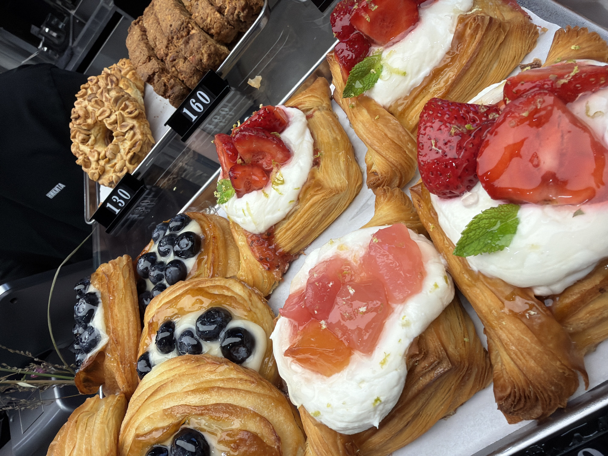 The image displays a close-up view of various pastries, likely in a bakery or cafe setting. The pastries are arranged on a metal tray. Several of them are danishes topped with white cream and garnished with strawberries, lime zest, and mint leaves. Others are danishes filled with blueberries and cream. There are also cookies visible in the background, including a ring-shaped cookie covered in nuts and some rougher, textured cookies.
The scene is well-lit, suggesting it could be daytime. There are price tags visible in front of some of the pastries. One tag reads "160" and another reads "130". The word "НИКИТА" is visible in white text on a dark background, likely on clothing, indicating a personal name or brand. The overall impression is one of a fresh display of baked goods.