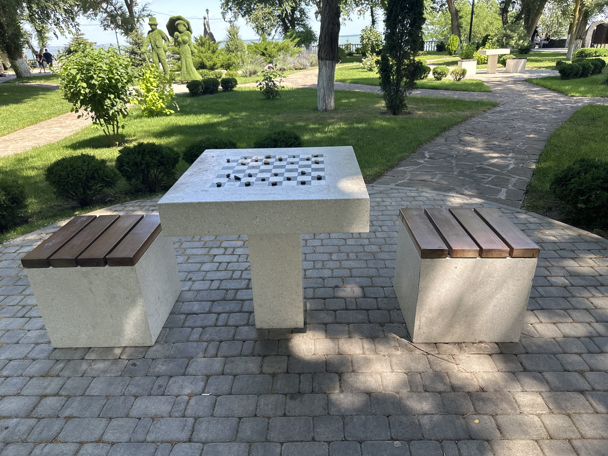 In Taman, Russia, a public park features a stone table with a built-in chessboard and two accompanying stone benches with wooden slats. The chessboard is set up with several stones, suggesting a game in progress. The table and benches are situated on a paved walkway made of interlocking gray stones. Surrounding the seating area is a well-maintained lawn with manicured bushes and trees. In the background, a pathway winds through the park, leading towards the sea, which is visible in the distance. Further in the background, two large topiary figures, one resembling a man and the other a woman, are prominent green sculptures. Several trees with visible trunks and foliage dot the landscape, and the scene is illuminated by natural sunlight casting shadows on the ground. A few people are visible in the distance on a pathway, walking.