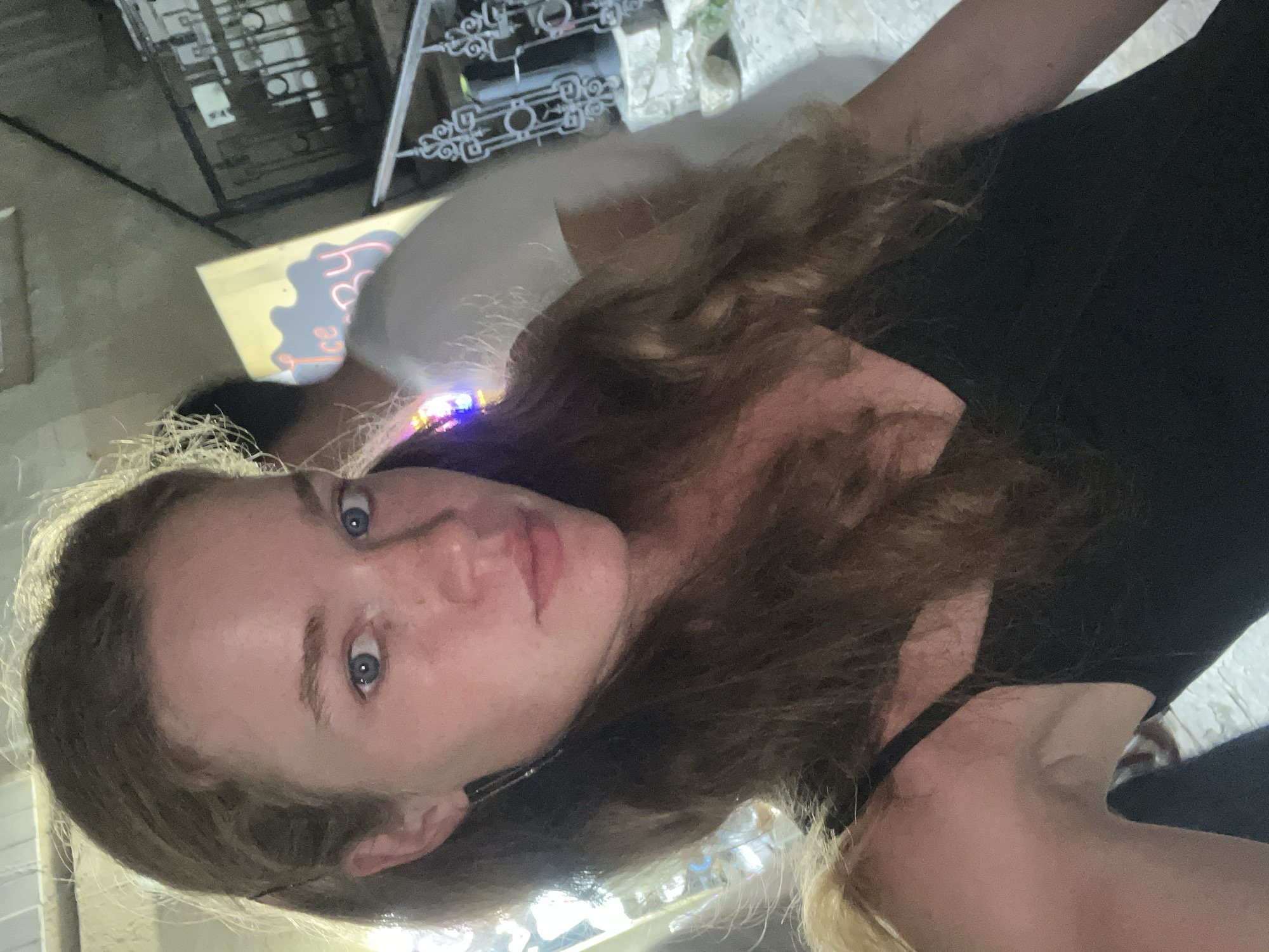 This media file shows a close-up, vertically oriented selfie of a young person. The subject has long, wavy brown hair and blue eyes. They are wearing a black tank top, and their skin appears fair with some freckles on their cheeks. The background is out of focus and dimly lit, suggesting an indoor setting, possibly at night. Hints of neon lights and some metallic structures are visible. The scene appears to be casual and personal. There is no discernible text in the image.