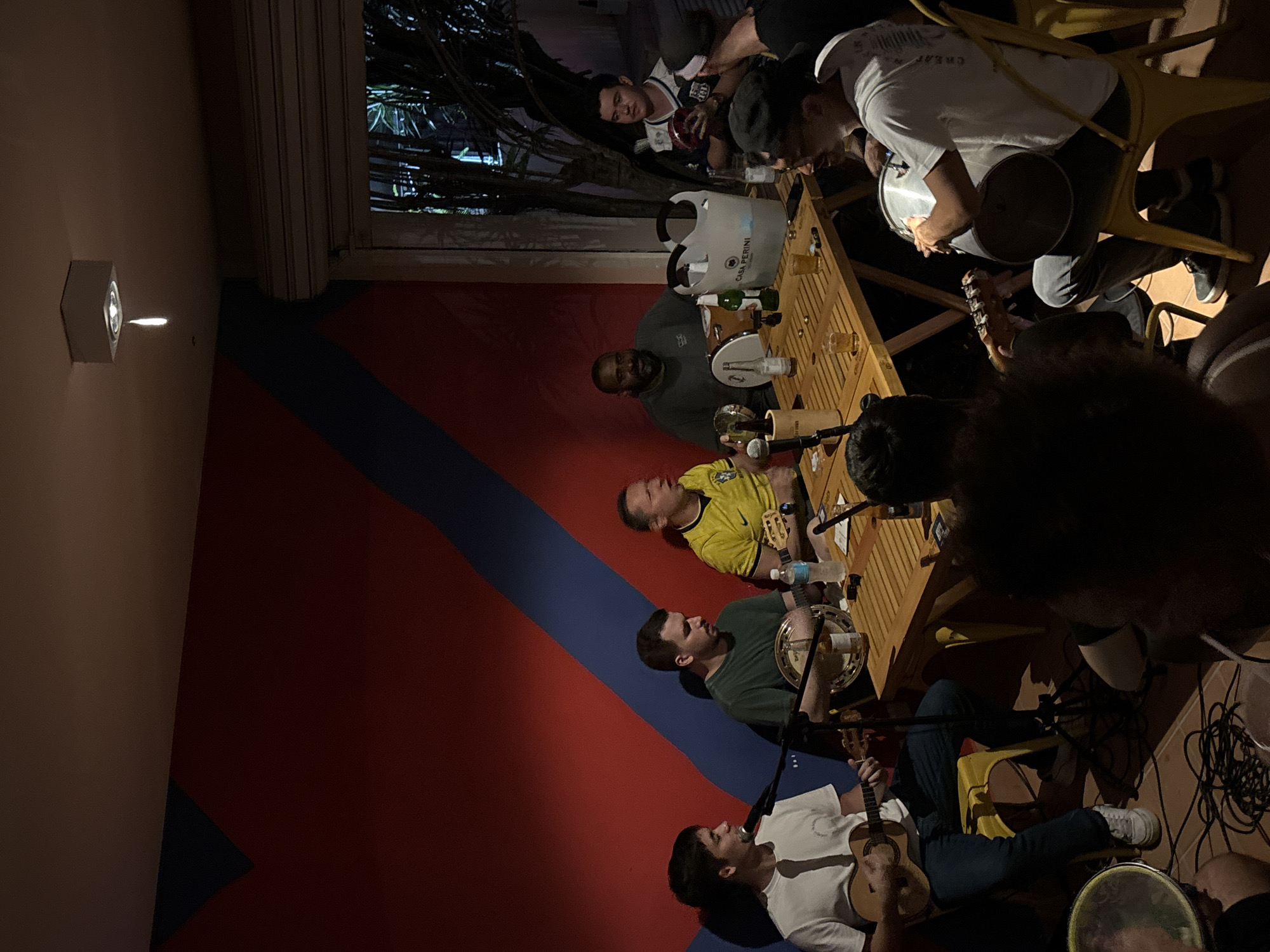 A group of men are gathered around a wooden table, some playing musical instruments. There are glasses and bottles on the table. A cooler labeled "Casa Perini" is visible. The scene is set indoors. The wall behind the group is painted in red and blue diagonal stripes. In the upper left corner, a window reveals vegetation outside. The location is São Paulo, Brazil.
