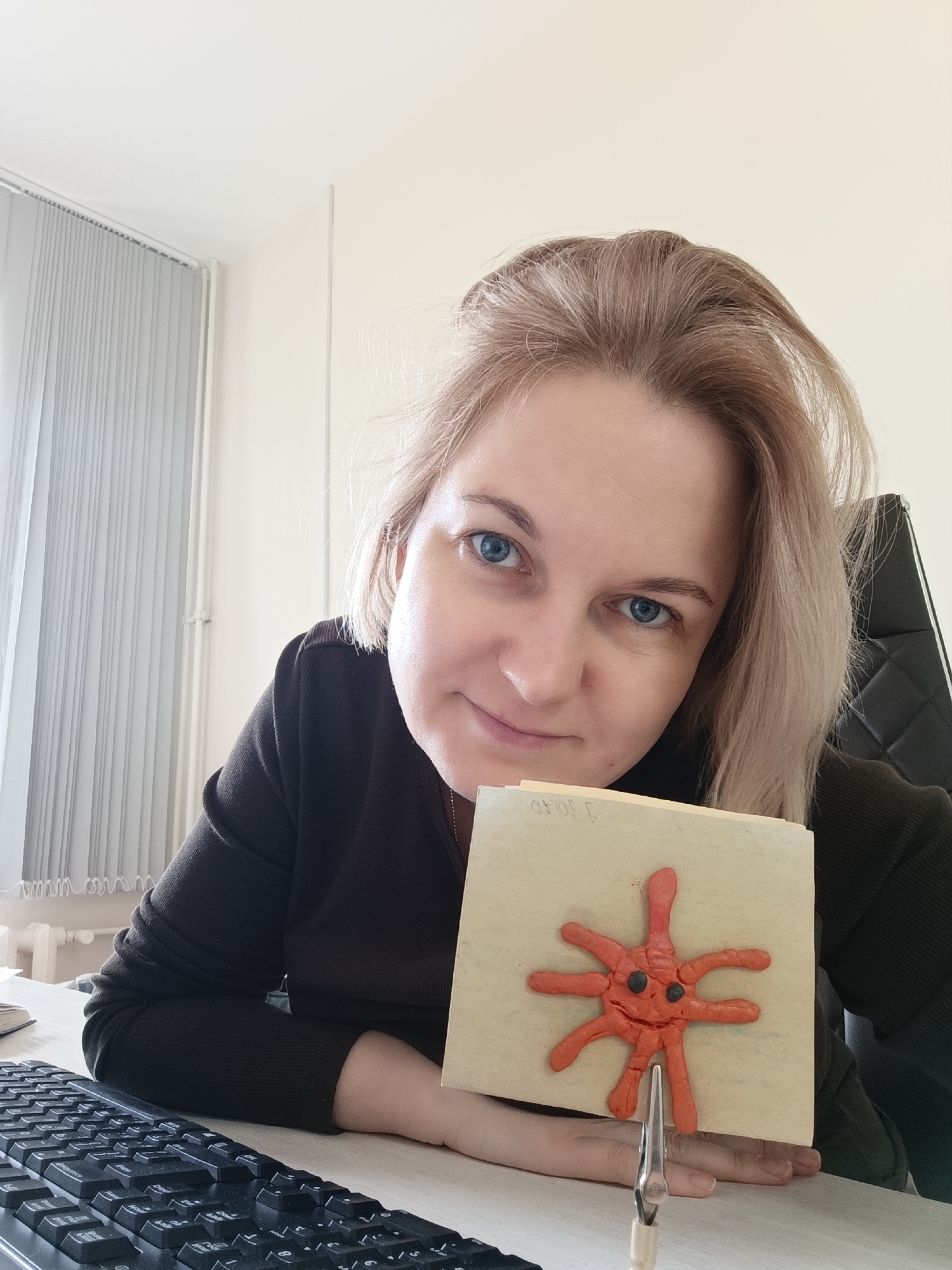 A woman with blonde hair and blue eyes is seated at a desk, holding up a square piece of beige paper with a clay figure of a stylized orange virus or bacteria attached to it. The clay figure has protruding tentacles and two black eyes. The paper is being held by a metal clip attached to its bottom edge. The woman is wearing a dark brown sweater. A black computer keyboard is visible on the desk in front of her. Behind her is a window with white blinds and a black office chair. The location is unspecified, but has been determined to be in the city of Markovskiy, Russia.