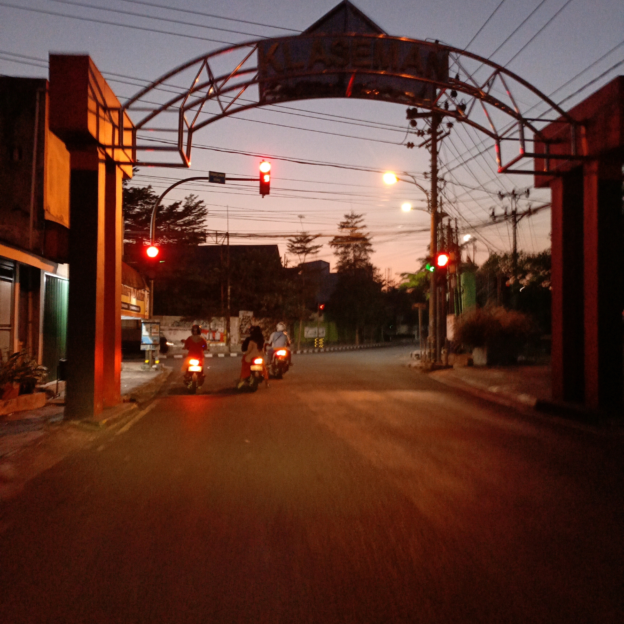 The image shows a street scene at dusk in Salatiga, Indonesia, under an archway with the word "KLASEMAN" visible. The sky is a dusky purple and orange. A traffic light with a red light illuminated is visible. Several people on motorcycles are riding down the street, their headlights and taillights creating streaks of light. The street is paved and appears wet, reflecting the lights. Buildings and trees line the sides of the street. The overall atmosphere is twilight, with artificial lights beginning to illuminate the scene.