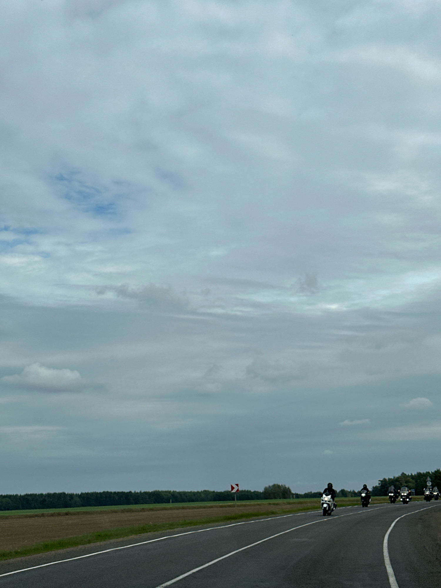 A group of motorcyclists ride in formation on a two-lane asphalt road. The road has white dashed and solid lines marking the lanes. To the left of the road is a flat, cultivated field, and beyond that, a dense line of trees forming a dark green horizon under a cloudy, overcast sky. The sky is filled with large, grey clouds with some lighter patches. A red and white directional sign is visible on the left side of the road. The motorcyclists are spaced out and appear to be traveling in the same direction, with at least five visible. They are wearing dark clothing and helmets. The overall scene suggests a travel or journey on a cloudy day in a rural area.
