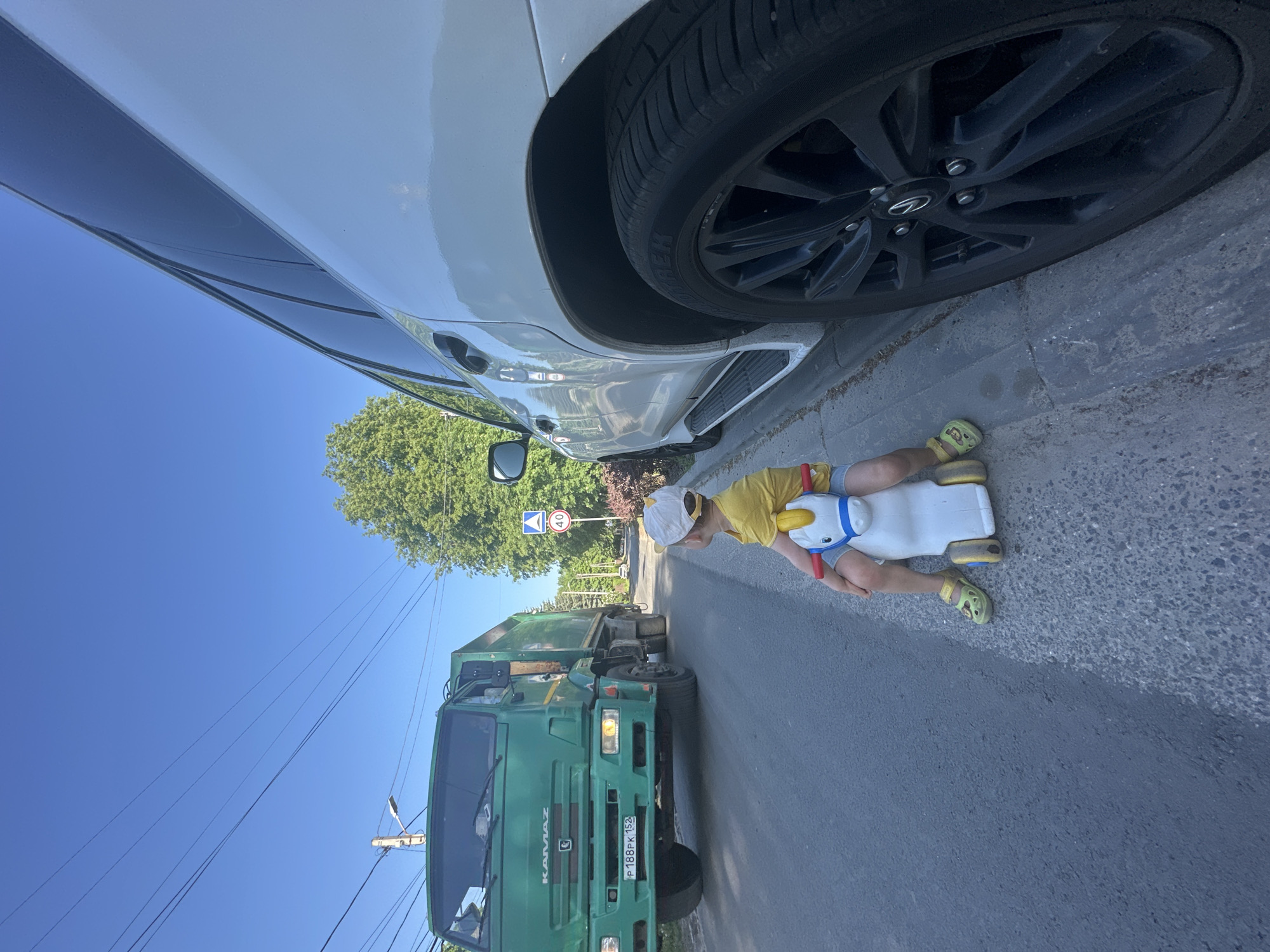 A low-angle, vertically oriented image depicts a scene on a sunny day, likely taken in Nizhny Novgorod, Russia, as indicated by the "KAMAZ" truck with a Russian license plate. A young child, dressed in a yellow shirt and shorts, lies prone on a white plastic ride-on toy on the asphalt road. The front passenger-side tire of a white SUV is positioned above and to the side of the child. In the background, a green KAMAZ truck approaches. A road sign indicating a speed limit of 40 km/h is visible further down the road. Overhead power lines extend across the blue sky.