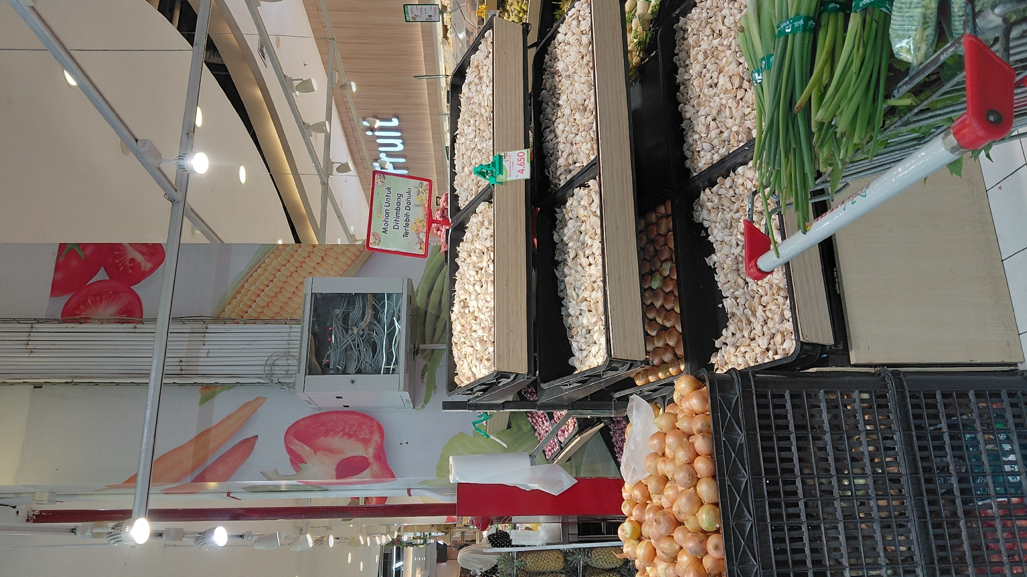 In a brightly lit supermarket produce section in Purwokerto, Indonesia, an assortment of fresh vegetables is displayed for sale. The foreground features a large black plastic crate filled with onions, with another crate of onions stacked behind it. To the right, several tiered black plastic display bins hold large quantities of peeled garlic cloves, with a price tag of 4.650 visible on one of the bins. Bunches of green onions are neatly arranged in a shopping cart in the lower right corner of the image, with the red handle of the cart extending into the frame. In the background, there is a large pillar decorated with vibrant imagery of vegetables such as tomatoes, carrots, and corn. A metal cabinet with visible wiring is mounted on the pillar. Further in the background, a sign with Indonesian text, likely indicating product information, is posted. The overall environment suggests a well-stocked grocery store catering to the local community.