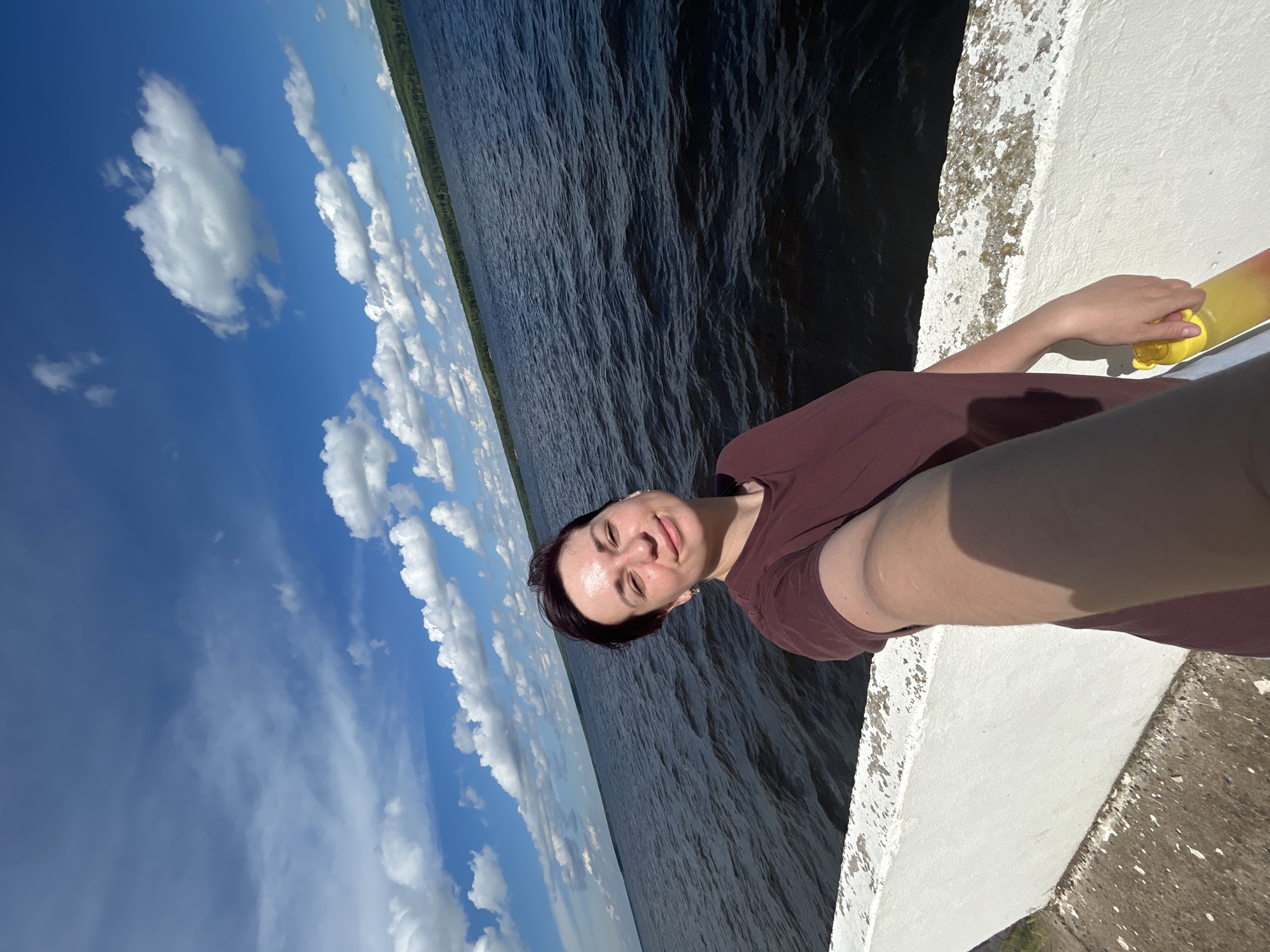 A woman with short, dark hair is taking a selfie outdoors. She is wearing a maroon t-shirt and is standing next to a white concrete barrier overlooking a dark, choppy body of water. The sky is blue with scattered white clouds, and a green, tree-lined shore is visible in the distance. The woman is holding a yellow water bottle in her right hand. The setting appears to be the embankment along the Kama River in Chaikovsky, Russia.