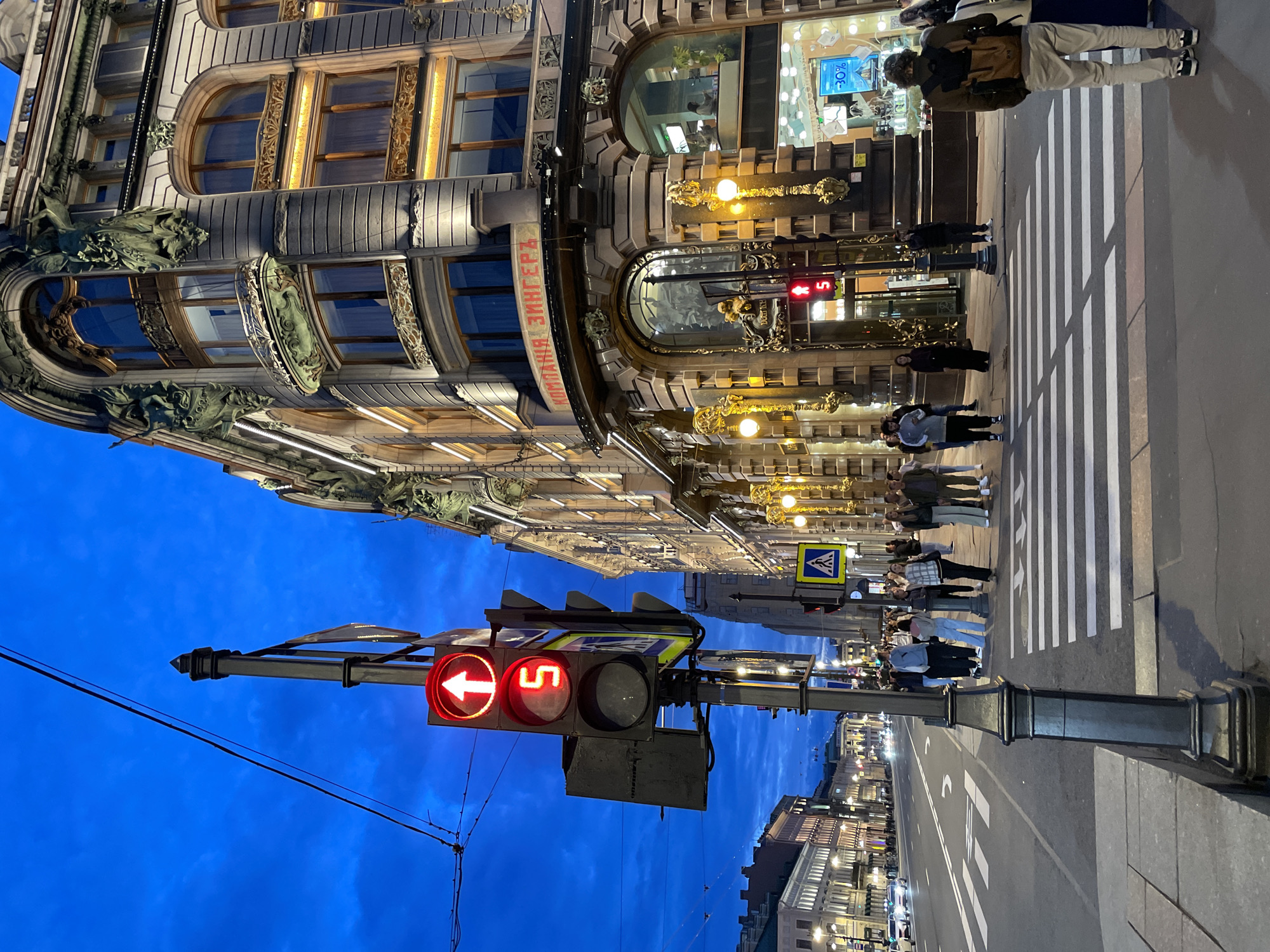 The image depicts an urban street scene at dusk. A traffic light with a red upward arrow and a red numeral "5" is prominently displayed in the foreground. The traffic light is mounted on a dark gray pole. Behind the traffic light, a grand, ornate building with multiple windows and architectural details is illuminated by interior lights and accent lighting along its facade. The building has signage with Cyrillic lettering. A pedestrian crossing is visible on the street, with several people waiting to cross or having just crossed. The street is wide and appears to be a main thoroughfare, with cars driving in the distance. The sky is a deep blue, indicative of twilight. The overall setting suggests a city environment with historical architecture.