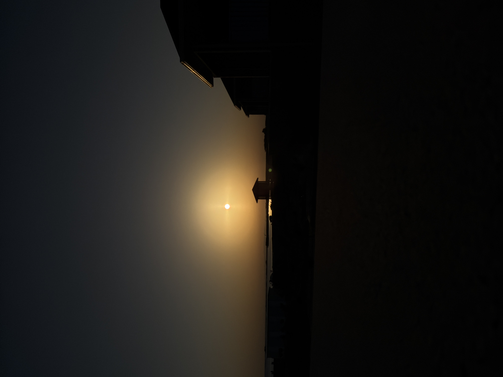 The image depicts a sunset over a body of water, likely the Caspian Sea, viewed from Pirallahi Raion, Azerbaijan. Silhouetted structures, including what appears to be a small building and larger buildings with angled roofs, are visible against the gradient of the sky. The sun, a bright orb, is near the horizon, casting a warm glow that transitions into a darker, dusky sky. The foreground is dark, obscuring details.