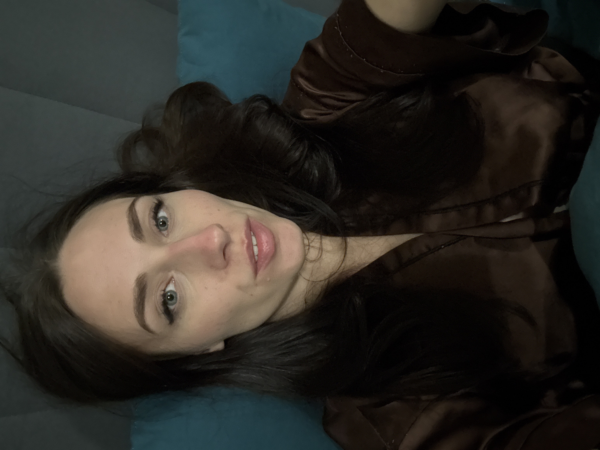 The image displays a close-up portrait of a young woman lying down, looking directly at the camera. Her head is tilted to the left, and her body is oriented horizontally within the vertically framed shot. She has long, dark brown hair that spreads out around her head, and her striking light blue or green eyes are open and focused. Her eyebrows are well-defined, and she wears light-colored lipstick on her slightly parted lips, revealing a hint of her teeth in a subtle, gentle smile.
She is dressed in what appears to be a dark brown or deep red satin-like robe or loungewear, visible draped around her upper body. The fabric has a soft sheen, suggesting a luxurious texture. The immediate background beneath her head and to her right is a teal or dark blue upholstered surface, likely a pillow or part of a bed or sofa. To her left, a darker, plain greyish surface is visible, possibly a wall or another piece of furniture.
The lighting is soft and diffused, indicating an indoor setting, possibly during the evening or night, given the ambient quality. The woman's expression is calm and direct, with a slight, inviting smile. There are no visible texts or specific environmental cues within the image to denote a particular location or weather conditions. While external context suggests Saint Petersburg, Russia, this is not discernible from the visual information alone.