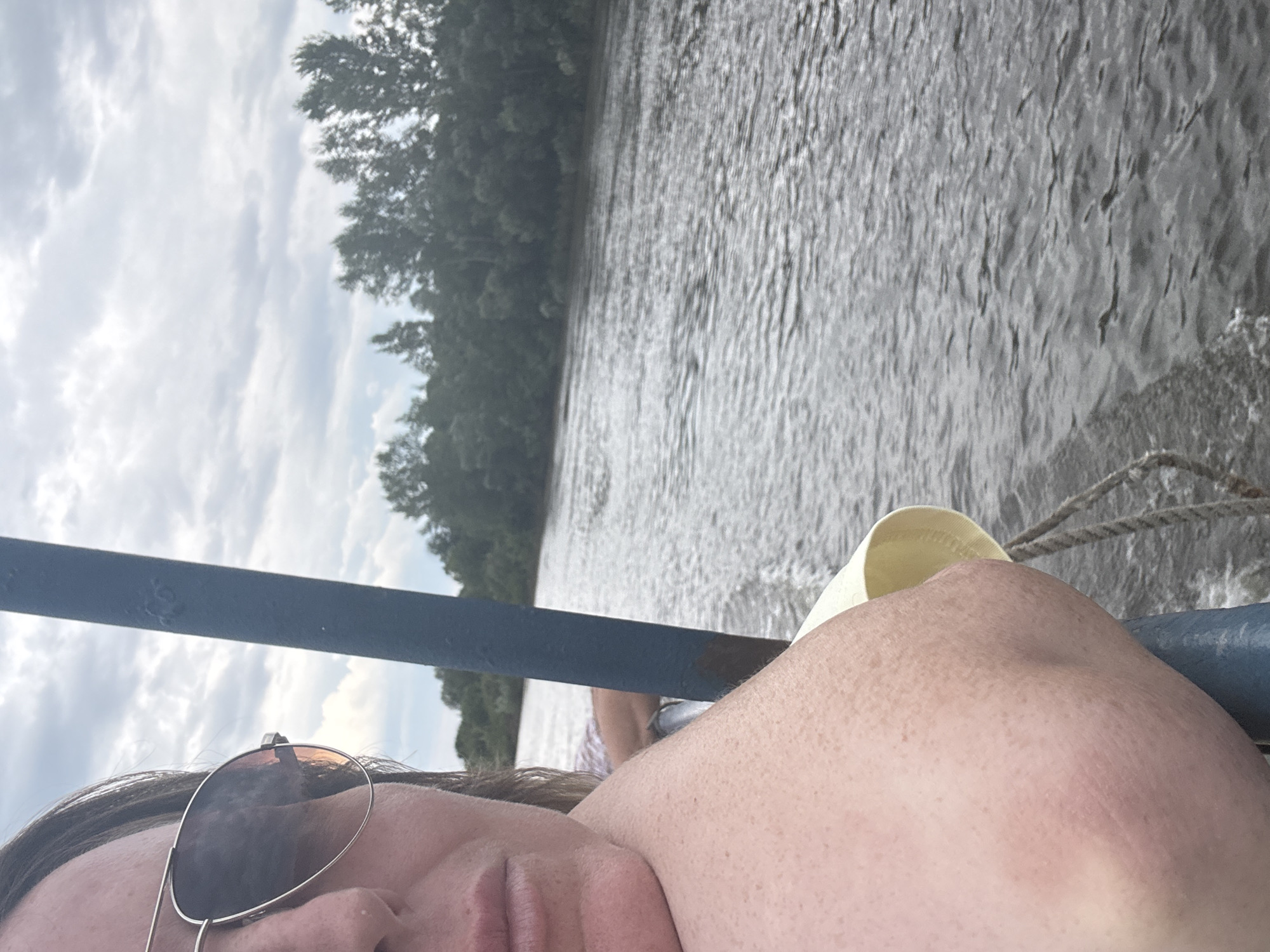 A woman wearing aviator sunglasses leans on a blue railing while on a boat. She has light-colored skin with freckles and is partially visible from the chest up, with her head turned to the side. The boat is on a wide, choppy river, with a forested bank visible in the distance under a cloudy sky. The scene appears to be in Ufa, Russia, but this is not explicitly stated in the image.