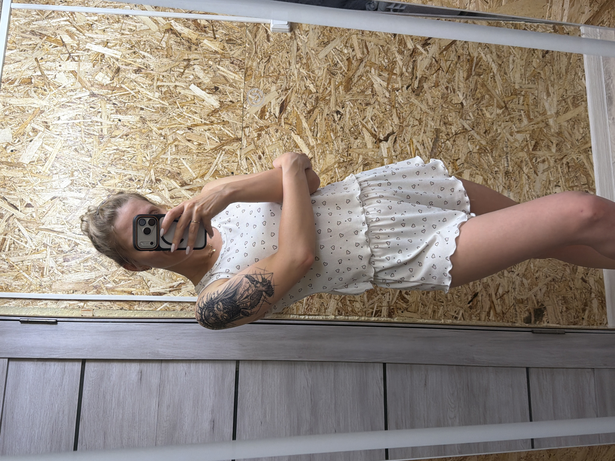 This full-body mirror selfie, taken in an indoor setting in the city of Chaikovsky, Russia, features a female-presenting individual. The person's face is largely obscured by a dark-colored smartphone, held in the right hand for the photograph, with its triple-lens camera module visible. The individual wears a white two-piece pajama set adorned with small dark heart patterns; the top is a tank top style with a ribbed hem, and the shorts have a matching ribbed and ruffled hem. Light-colored nail polish is visible on the fingernails of the hand holding the phone, and a ring is present on the right ring finger. A delicate gold-colored necklace with a small pendant is visible on the upper chest. The individual has light-colored hair styled in an updo and a prominent, intricate black-ink tattoo on the left upper arm, depicting a figure with a crowned or spiked head, a partially obscured face, and two birds below. The background, as reflected in the mirror, consists of two distinct sections: to the left, a light-colored wood-paneled wall or door with horizontal dark lines and a visible silver door hinge; to the right, a wall covered in oriented strand board (OSB) with its characteristic irregular wood chip texture. A light-colored floor is visible at the bottom of the image.