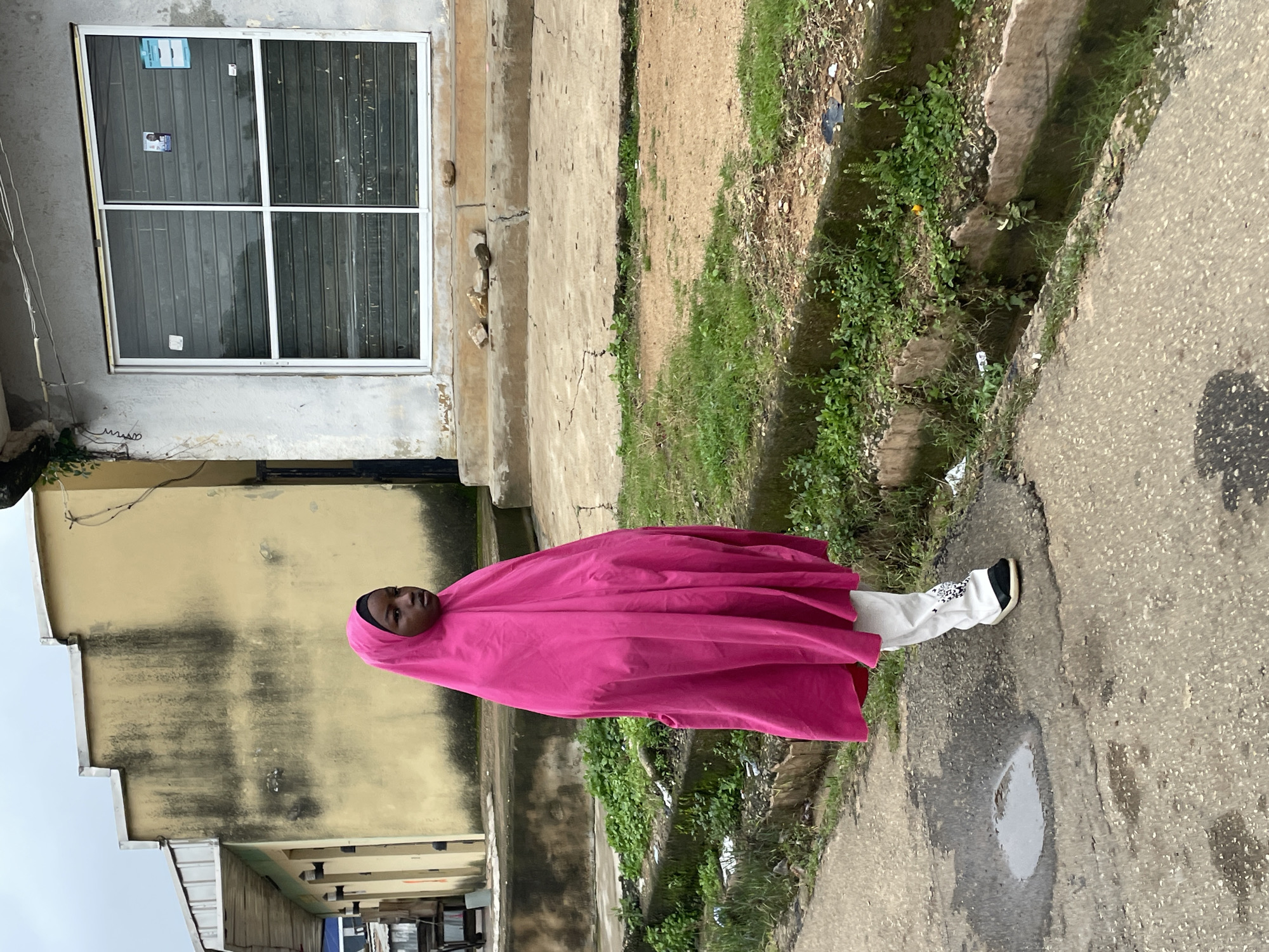 In Jos, Nigeria, a young person wearing a bright pink hijab and white pants stands on an overcast day. The individual is positioned on a paved road with a large puddle to their left and a concrete drainage channel filled with green vegetation to their right. Behind them, a pale yellow building with dark stains runs parallel to the road, featuring several outdoor light fixtures. Further back and to the right, a grey building with a large, shuttered window is visible. The overall scene suggests a typical urban environment in Jos, with a focus on the pedestrian and the surrounding infrastructure.
