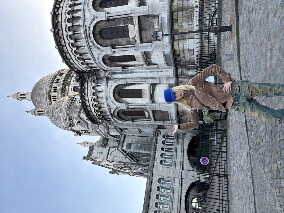 A female individual stands on a cobblestone surface in front of the Sacré-Cœur Basilica in Montmartre, Paris, France. She has blonde hair and wears a blue cylindrical hat, a brown long-sleeved jacket with a diagonal button closure, a black top partially visible at the waist, and severely distressed light blue jeans. Her left arm is raised with an open palm, while her right hand rests on her hip. She faces the camera directly. Behind her is a black metal fence, a lamppost, and a "No Parking" sign. The grand white stone facade and domes of the Sacré-Cœur Basilica rise prominently in the background under a clear sky.