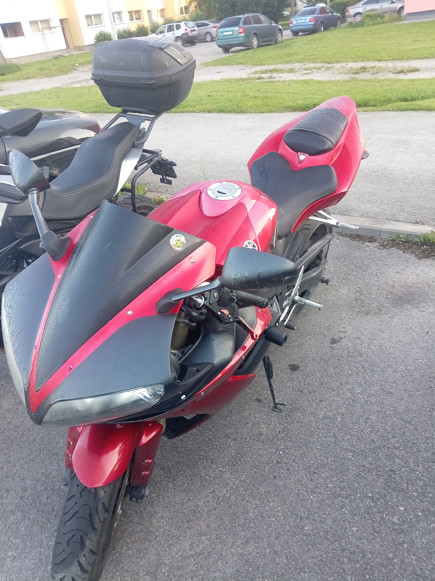 A red motorcycle is parked on an asphalt surface. The motorcycle has a black windscreen, black seats, and a black storage box attached to the rear. The motorcycle is wet, indicating it may have rained recently. In the background, there are several cars parked and a building. The location is the city of Tapa, Estonia.