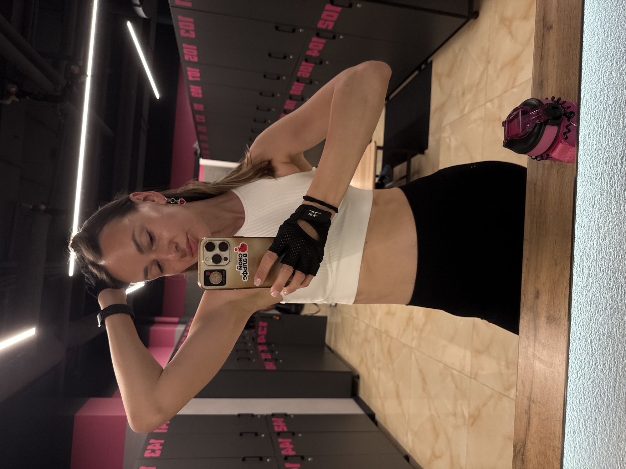 A woman with her hair pulled back is taking a selfie in a gym locker room. She is wearing a white crop top, black leggings, and black fingerless gloves. Her left arm is raised, and she is touching her head. Her right hand is holding a smartphone, which is showing a picture of herself with stickers. She is wearing red heart-shaped earrings and a black watch on her left wrist. The locker room has gray lockers with pink lettering and a beige tiled floor. A wooden bench and a pink water bottle are visible in the reflection of a mirror. The image appears to have been taken in Sochi, Russia.