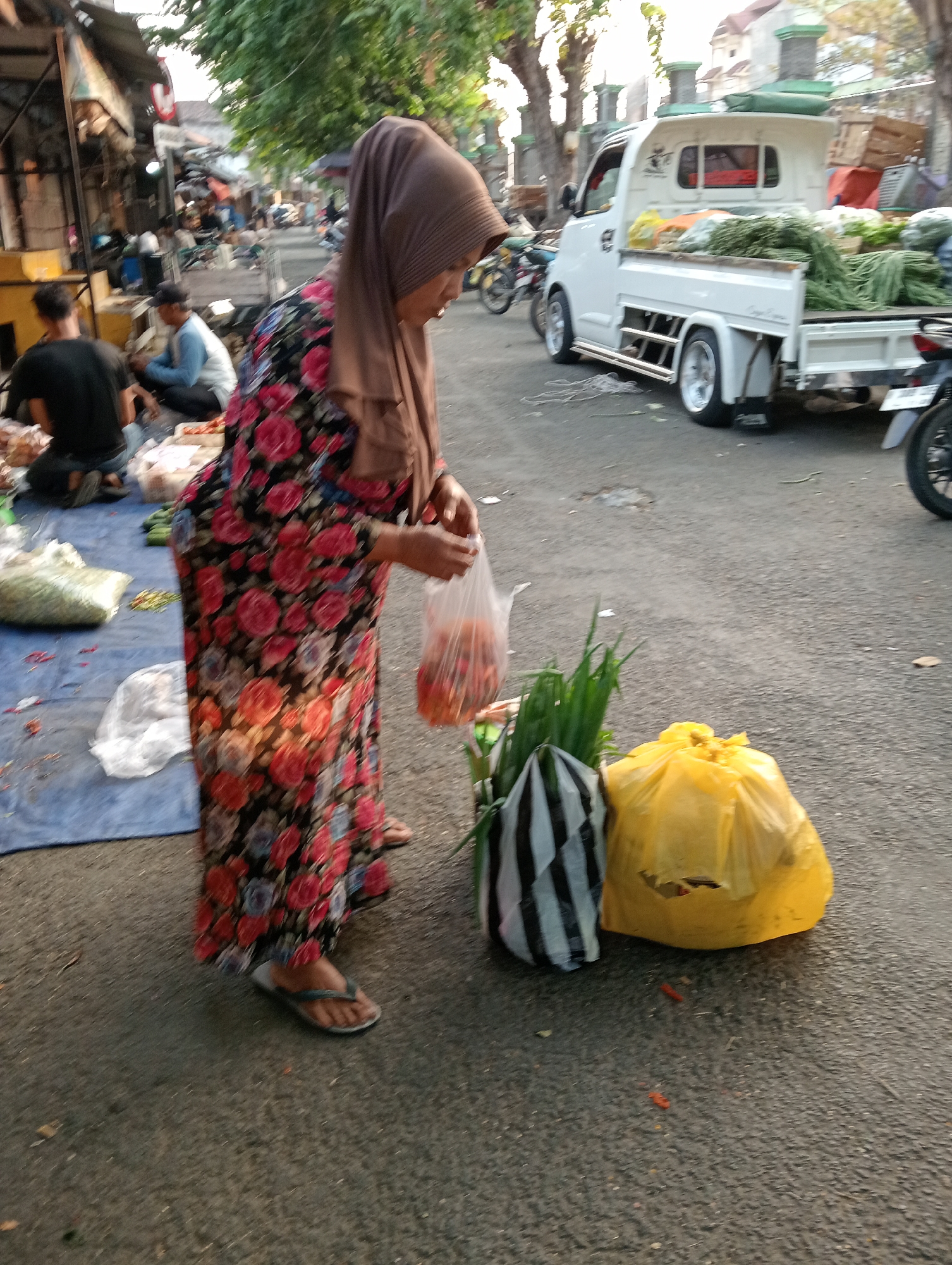 In Demak, Indonesia, a woman wearing a brown hijab and a floral-patterned dress stands on an asphalt road, holding a plastic bag filled with red vegetables. To her right, a white pickup truck is loaded with produce, including green beans. In the foreground, there are two plastic bags, one with black and white stripes containing green onions, and another yellow plastic bag. Other people are visible in the background, as well as market stalls and motorcycles. The scene appears to be a street market.
