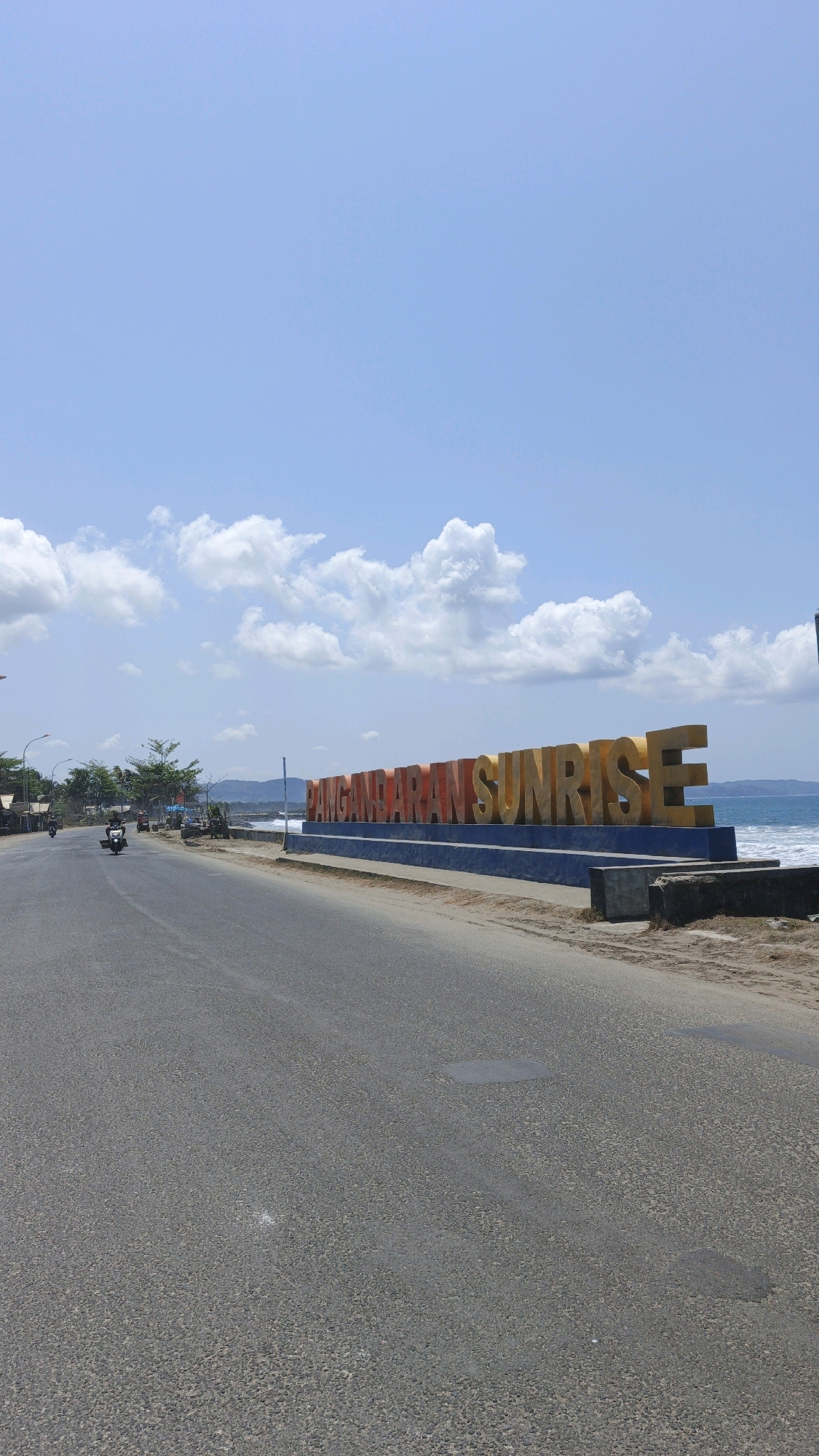 An eye-level, vertical, medium shot captures the "PANGANDARAN SUNRISE" monument situated alongside a road on a bright, sunny day in Desa Pangandaran, Indonesia. The monument, composed of large, three-dimensional letters in orange and yellow, stands on a blue platform. The letters spell out "PANGANDARAN SUNRISE" and are illuminated by sunlight, creating shadows on the ground.
To the left of the monument, a two-lane asphalt road stretches into the distance, with a slight curve to the left. The road is dry and shows some wear and tear. On the left side of the road, there is a sandy shoulder with scattered vegetation, including palm trees, and several buildings with reddish-brown roofs, indicative of a coastal town. A few motorcycles are visible on the road and sidewalk. One motorcycle, ridden by an individual, is in motion, heading away from the camera.
The background features the Indian Ocean, with gentle waves lapping at the shore. The sea is a deep blue, transitioning to a lighter blue towards the horizon. A few distant hills or mountains are visible against the skyline. The sky above is a clear, bright blue, dotted with scattered white, puffy cumulus clouds. The overall scene depicts a roadside attraction in a coastal environment, under a clear, sunny sky.