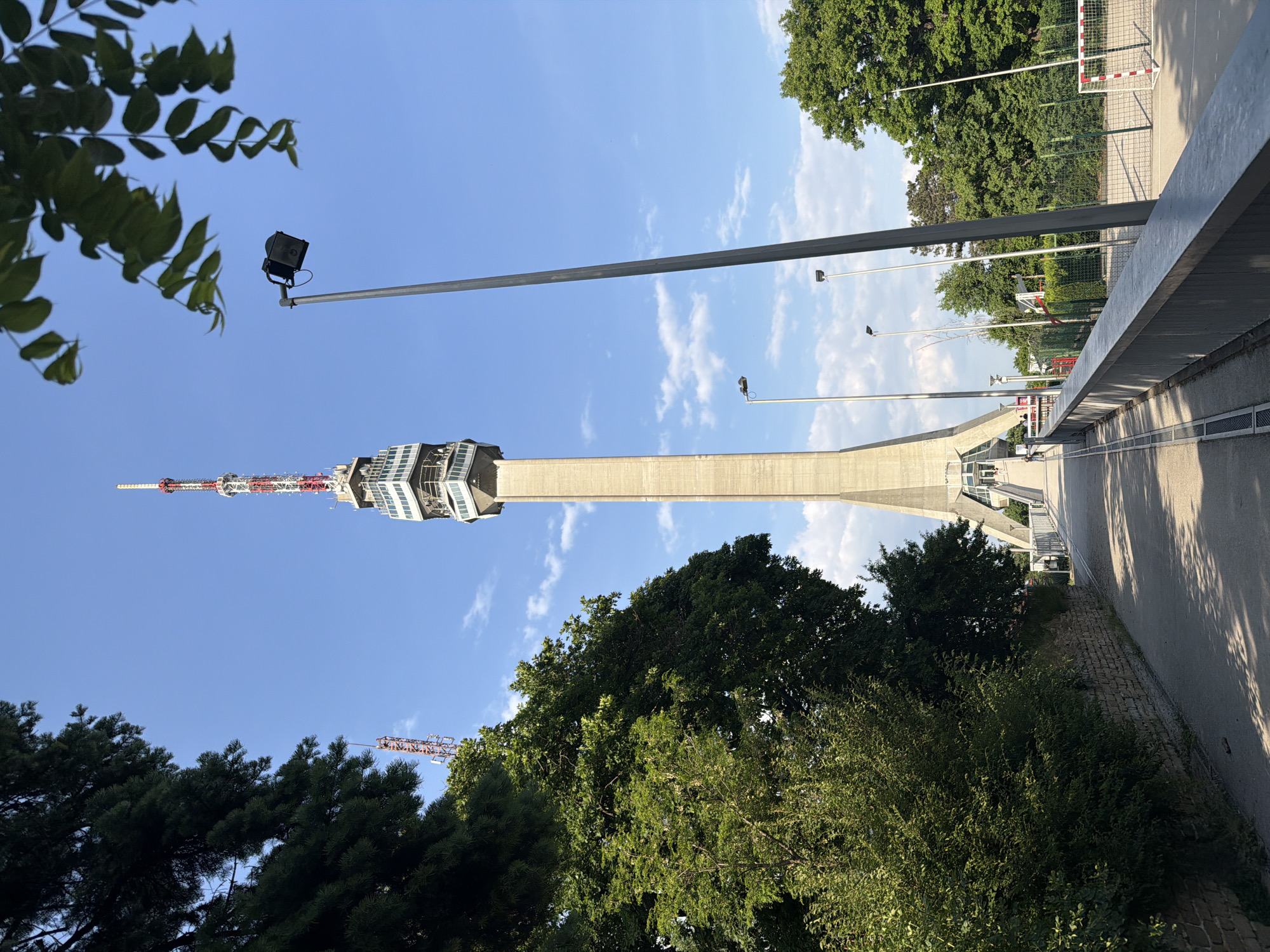 The image shows the Avala Tower in Vozdovac Urban Municipality, Serbia, viewed from a ground-level perspective, looking upwards. The concrete tower structure, with its distinctive observation deck and antenna, dominates the composition against a blue sky with scattered clouds. Trees frame the view, both at the bottom and along the top right. A walkway with a gray railing is visible to the right of the tower. Several light poles are positioned along the walkway. A soccer goal net is visible in the background, behind the trees on the right. A construction crane is partially visible in the trees. The scene is bathed in sunlight, suggesting a clear day.