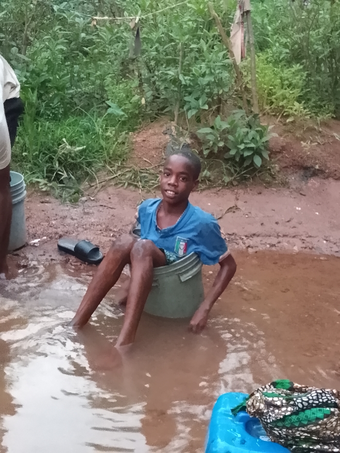 A young, smiling boy sits in a green bucket, within a pool of muddy water. He wears a blue t-shirt with the Italian flag emblem. To his left, a black sandal sits on the muddy ground. Behind him, lush green foliage is visible. To the right, a blue plastic container is partially submerged in the water, with a patterned cloth draped over it. A person's legs and a gray bucket can be seen to the left of the image.