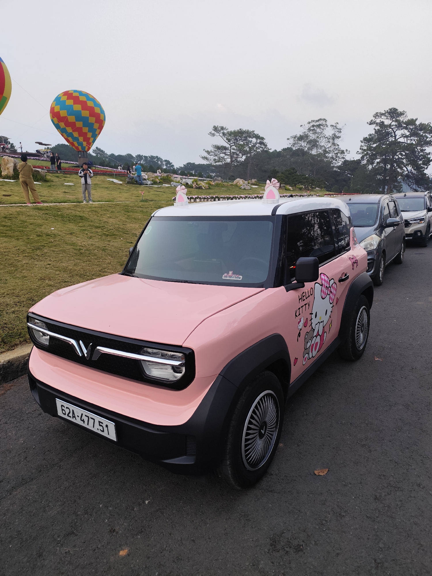 The image depicts a vibrant outdoor scene in Da Lat, Vietnam, likely within a tourist park such as Thung Lũng Tình Yêu (Valley of Love). The foreground is dominated by a pink and white compact electric vehicle, identifiable as a VinFast VF 3. The car features "Hello Kitty" themed graphics on its side, illustrating Hello Kitty, a small bear, and hearts. Pink cat ear accessories are affixed to the front roof, and a pink bow ornament is visible on the rear roofline. The vehicle displays a Vietnamese license plate, "62A-477.51", on its front bumper. Behind this car, a line of dark-colored passenger vehicles is parked on the asphalt road.
To the left and rear of the vehicles, a grassy slope ascends, populated by several individuals. One person in a light brown jumpsuit stands facing away from the camera, while another in a white jacket and dark pants is visible holding a mobile device. In the mid-ground, two large, tethered hot air balloons are partially inflated. One balloon exhibits a pattern of red and yellow panels, and the other a checkerboard design of blue, yellow, and red. The landscape includes landscaped grassy areas, decorative white animal figures (one adorned with a pink bow), and distant pine trees beneath an overcast sky.