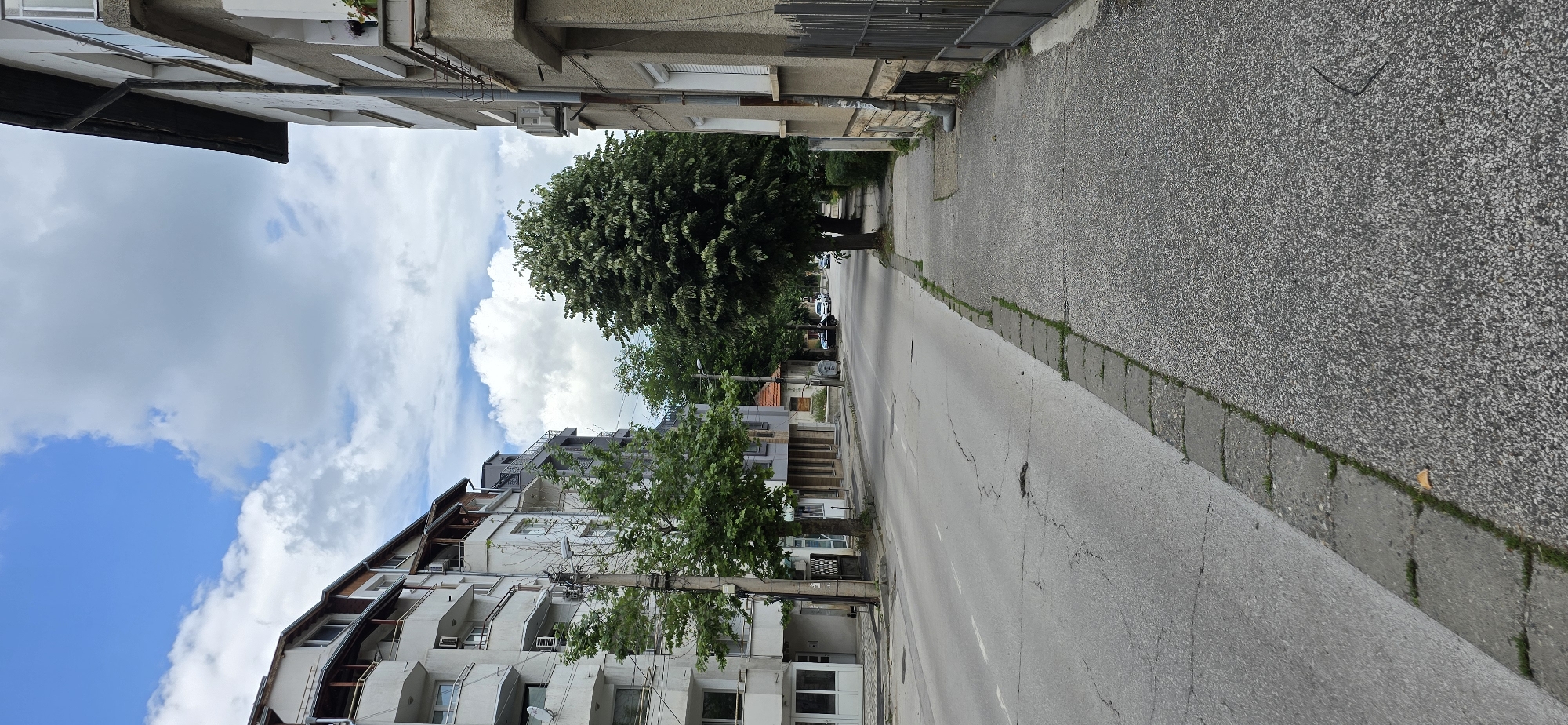 This image captures a street scene in the city of Unknown, Unknown, under a partly cloudy sky with patches of blue. The perspective is from the sidewalk, looking down the street.
To the left, a multi-story building with a light-colored facade, likely an apartment complex, features multiple balconies with mixed concrete and wooden railings. Utility conduits are visible on its exterior. A ground-level entrance with glass doors is at the far left. Further down the street on this side, other buildings with varying architectural styles and colors are partially visible. A utility pole with multiple overhead wires stands in the foreground on the left.
To the right, a multi-story building with a light brown textured facade is visible. It features windows, balconies, and an exposed dark roof edge. An external air conditioning unit is mounted on its facade. A vertical downspout runs along the building's exterior. A dark grey metal gate is visible in the lower right, providing access to the property.
The asphalt road, which is the central feature, displays numerous cracks, patches, and signs of wear. A faded white line runs longitudinally down the center of the visible road section. Several parked vehicles are discernible further down the street on both sides, suggesting a residential or local thoroughfare.
The sidewalk in the foreground, on the right side, is composed of gray aggregate concrete slabs, showing some surface texture. It is bordered by a curb made of rectangular grey pavers, with grass and small weeds growing in the gaps between the pavers and along the edge of the asphalt. A small, light brown leaf rests on the curb near the bottom center. A dark, thin, curved object (possibly a wire or piece of debris) lies on the sidewalk in the lower right corner.
Numerous deciduous trees with full green foliage line both sides of the street, providing shade and obscuring some of the buildings further in the distance. A particularly large, dense tree stands prominently on the right side of the street, centrally within the frame, with its canopy extending wide. No individuals are present in the visible scene.