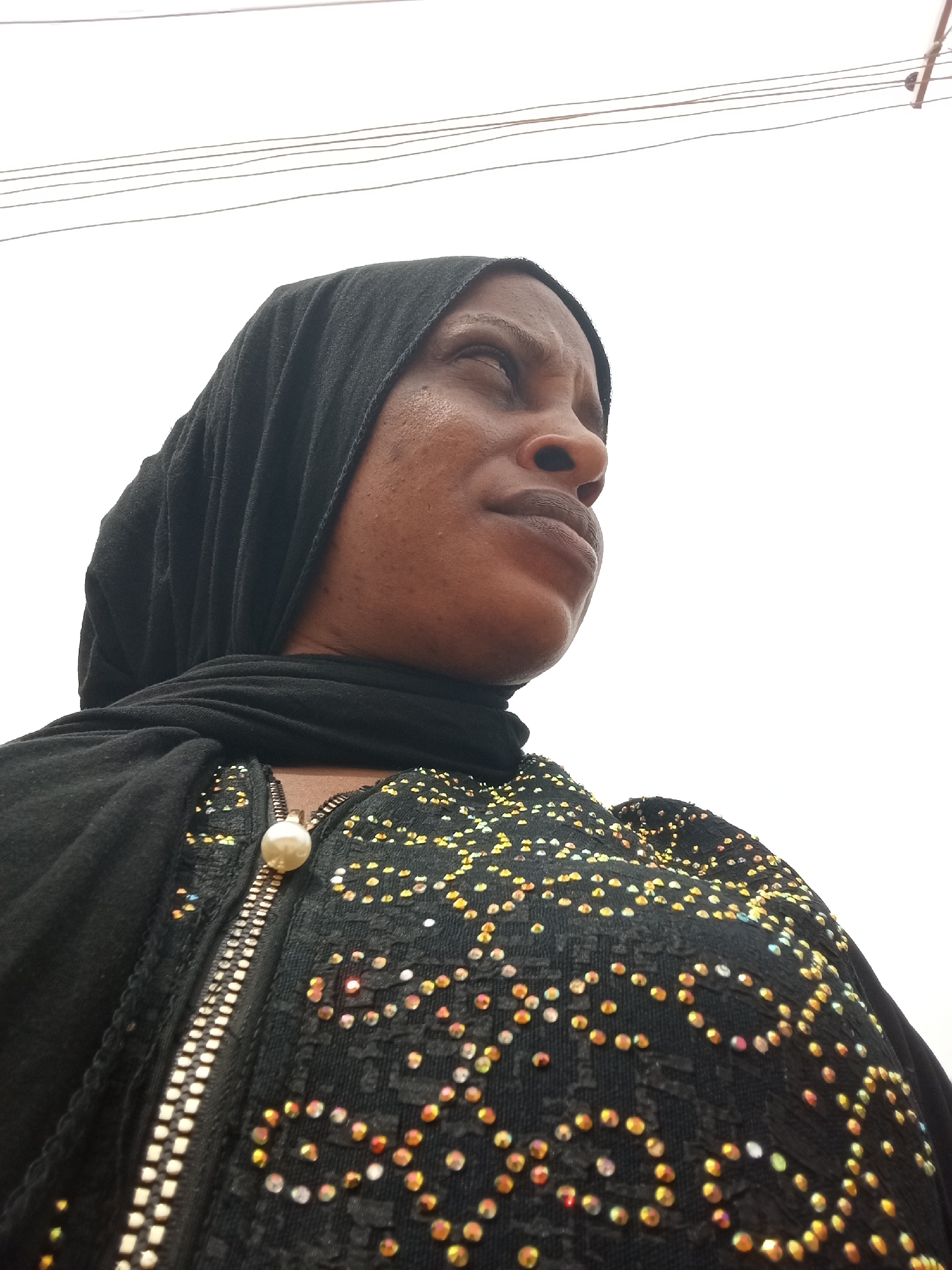 The image is a low-angle, medium shot of a woman in Narayi, Nigeria. She is wearing a black hijab and a black top or jacket adorned with a pattern of yellow and red rhinestones. The jacket has a silver zipper with a pearl-like pull tab. Overhead, there are several thin wires spanning the top of the frame against a bright, overcast sky. The woman is looking upwards and towards the right side of the frame. The camera appears to be held below her eye level.