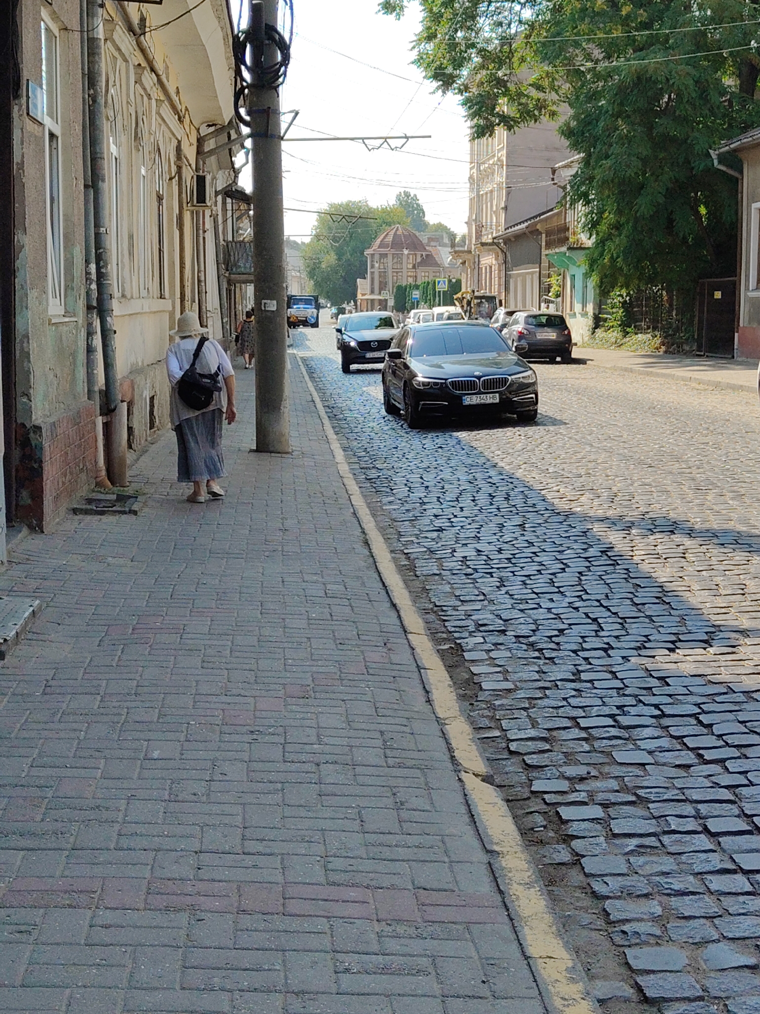 A woman wearing a hat and a long skirt walks down a cobblestone street in Chernivtsi, Ukraine. She is carrying a black bag over her shoulder. Cars are parked and driving on the street. Buildings line the left side of the street, and trees and more buildings are visible on the right. The sun is shining, casting shadows on the cobblestones. The street appears to be an older part of the city, with historic architecture.