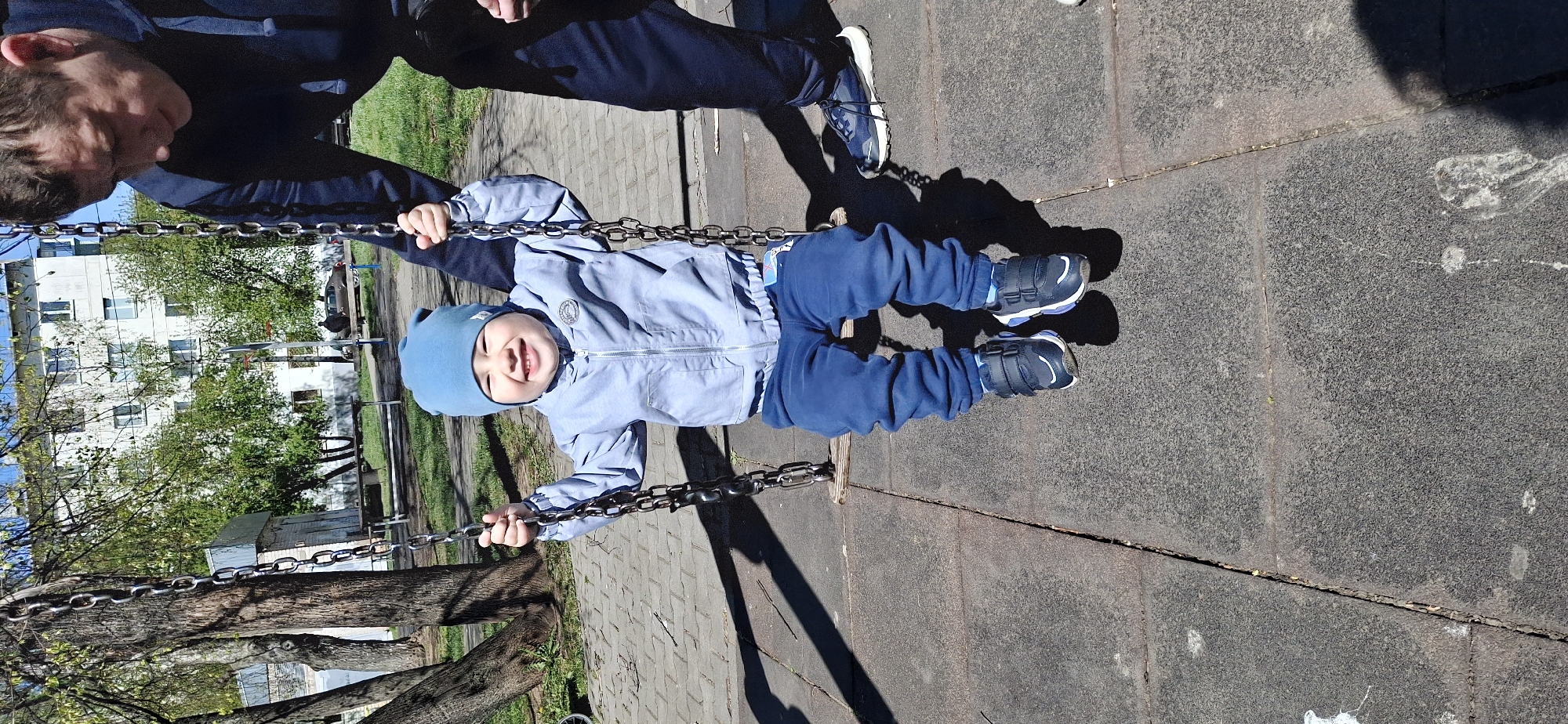 A young child is being pushed on a swing by an adult in the city of Chaikovsky, Russia. The child, wearing a blue hat, light blue jacket, and dark blue pants and shoes, is smiling. The adult is wearing dark clothing and dark shoes. The swing is made of metal chains. The ground is paved with grey blocks. Trees and a white building are visible in the background.