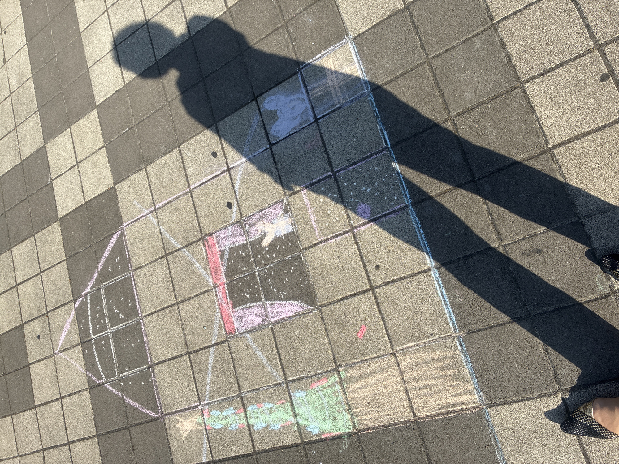 This is an eye-level, medium shot of a chalk drawing on a tiled walkway. The drawing depicts a stylized house with a triangular roof, a door, and a chimney. There is also a chalk drawing of a Christmas tree with an ornament to the left of the house. A shadow of a person is cast over the drawing, obscuring parts of it. The walkway is made of alternating light and dark gray square tiles. The lighting suggests it is daytime, with sunlight creating distinct shadows. The scene is outdoors, on a pedestrian walkway in the city of Sochi, Russia.