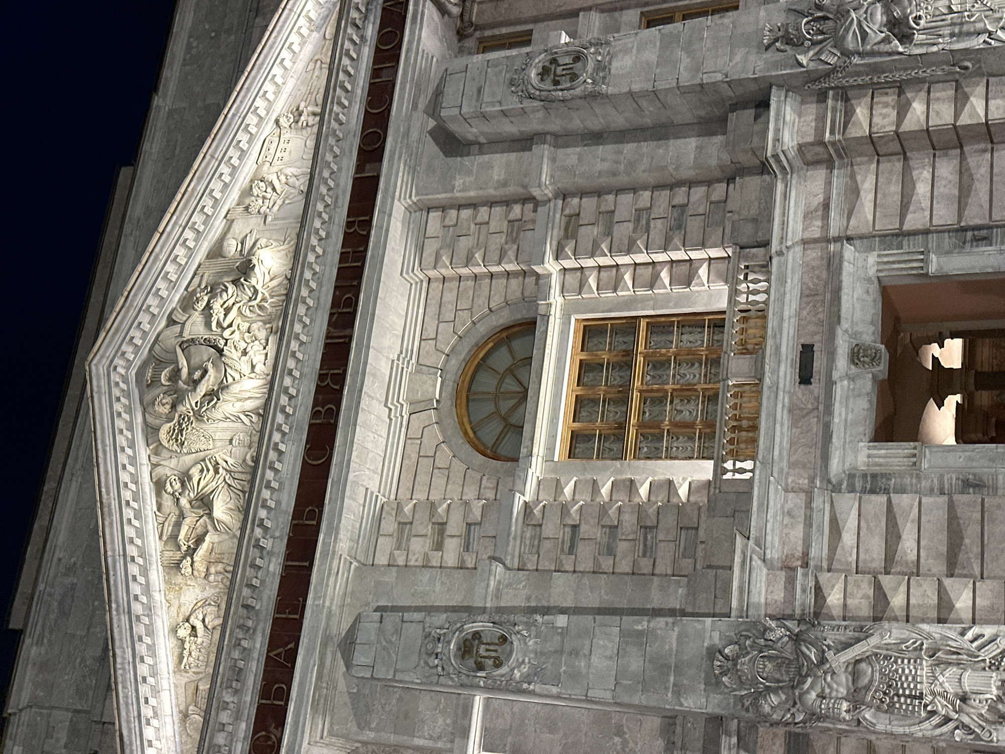 This is a nighttime, low-angle, medium shot of the facade of the Mikhailovsky Palace, also known as the Russian Museum, located in Saint Petersburg, Russia. The image focuses on the upper portion of the building, showcasing intricate architectural details.
The pediment of the building features a detailed bas-relief sculpture depicting allegorical figures. Below the pediment, a dark red band runs across the facade, inscribed with Cyrillic lettering. The main part of the facade is constructed from large, light-colored stone blocks. A prominent arched window with a wooden frame is visible, set above a wooden-paned rectangular window that has a small balcony with a decorative railing in front of it. On either side of the facade, flanking the central section, are vertical stone pilasters. The left pilaster has an oval medallion with a coat of arms, and the right pilaster has a similar medallion. Further down on the left and right sides, there are large, detailed sculptures of imposing, armored figures, each holding a weapon. The overall impression is one of grandeur and historical significance. The lighting suggests artificial illumination, casting shadows and highlighting the architectural features.