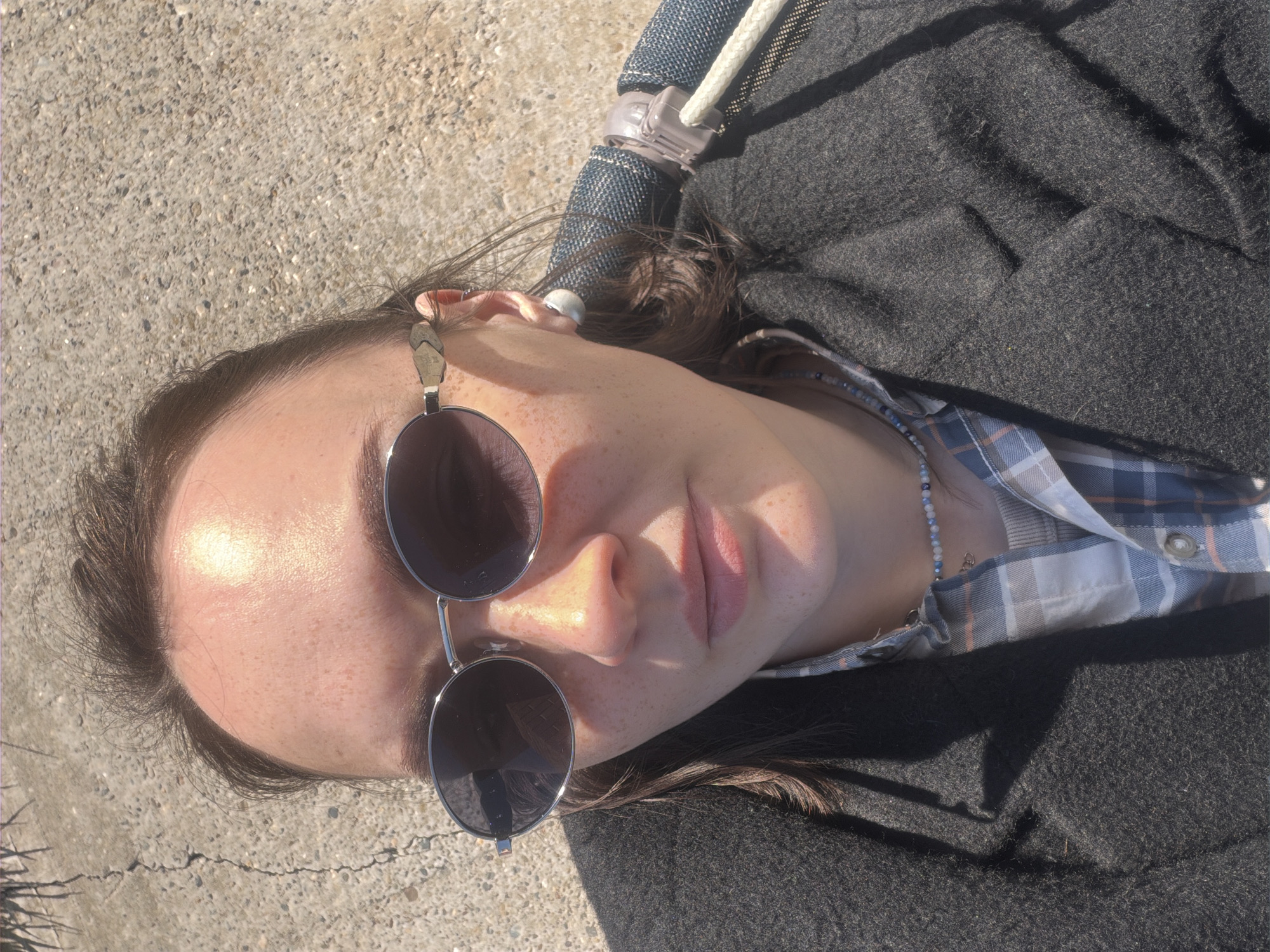 An overhead shot shows a person lying on what appears to be a concrete surface. The person is wearing sunglasses, a plaid shirt under a dark grey jacket, and a beaded necklace. Part of a denim hammock with a white rope support is visible above the person. The location is not discernible from the image and there is no way to identify Prikamskii, Russia as the location.