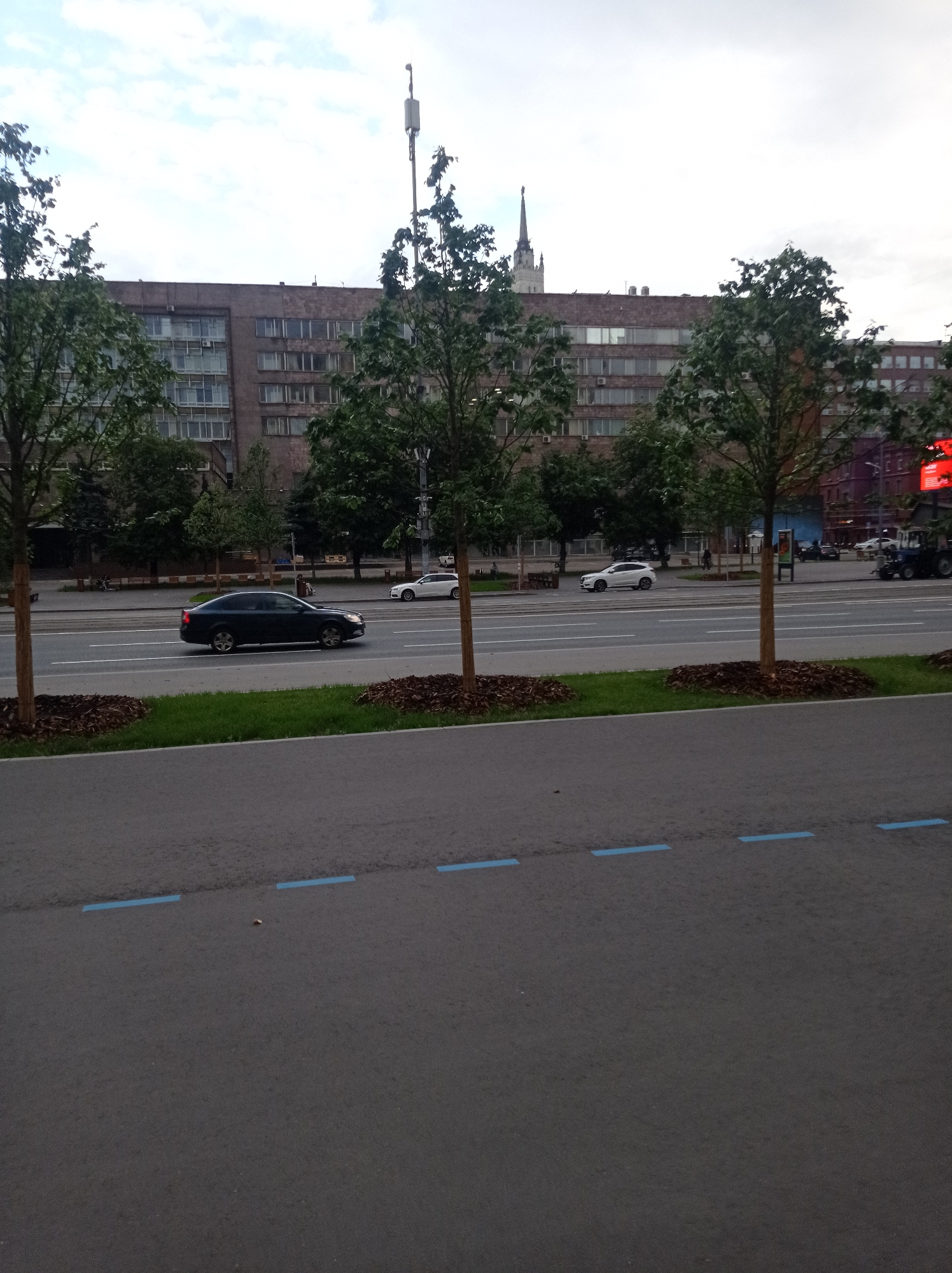 The image depicts a street scene in Moscow, Russia, likely during the daytime given the illumination and the presence of clouds. The foreground shows a paved road with several blue painted lines, suggesting it might be a bike lane or a special parking zone. Beyond the road, there's a grassy area with young trees planted along the edge.
In the middle ground, a multi-lane road is visible with several cars driving. A dark sedan is in the left lane, and a white SUV is in the right lane, suggesting moderate traffic. The road is bordered by a sidewalk or pedestrian area with benches, and more trees.
The background is dominated by a large, rectangular building with numerous windows, characteristic of Soviet-era architecture. A tall antenna tower is positioned on top of this building. To the right of the building, a tall, ornate spire can be seen, possibly part of a church or another significant landmark, which helps place the location within Moscow. The sky is overcast with clouds, indicating a potentially cool or temperate day. There are no people clearly visible and identifiable, nor any discernible text in the image.