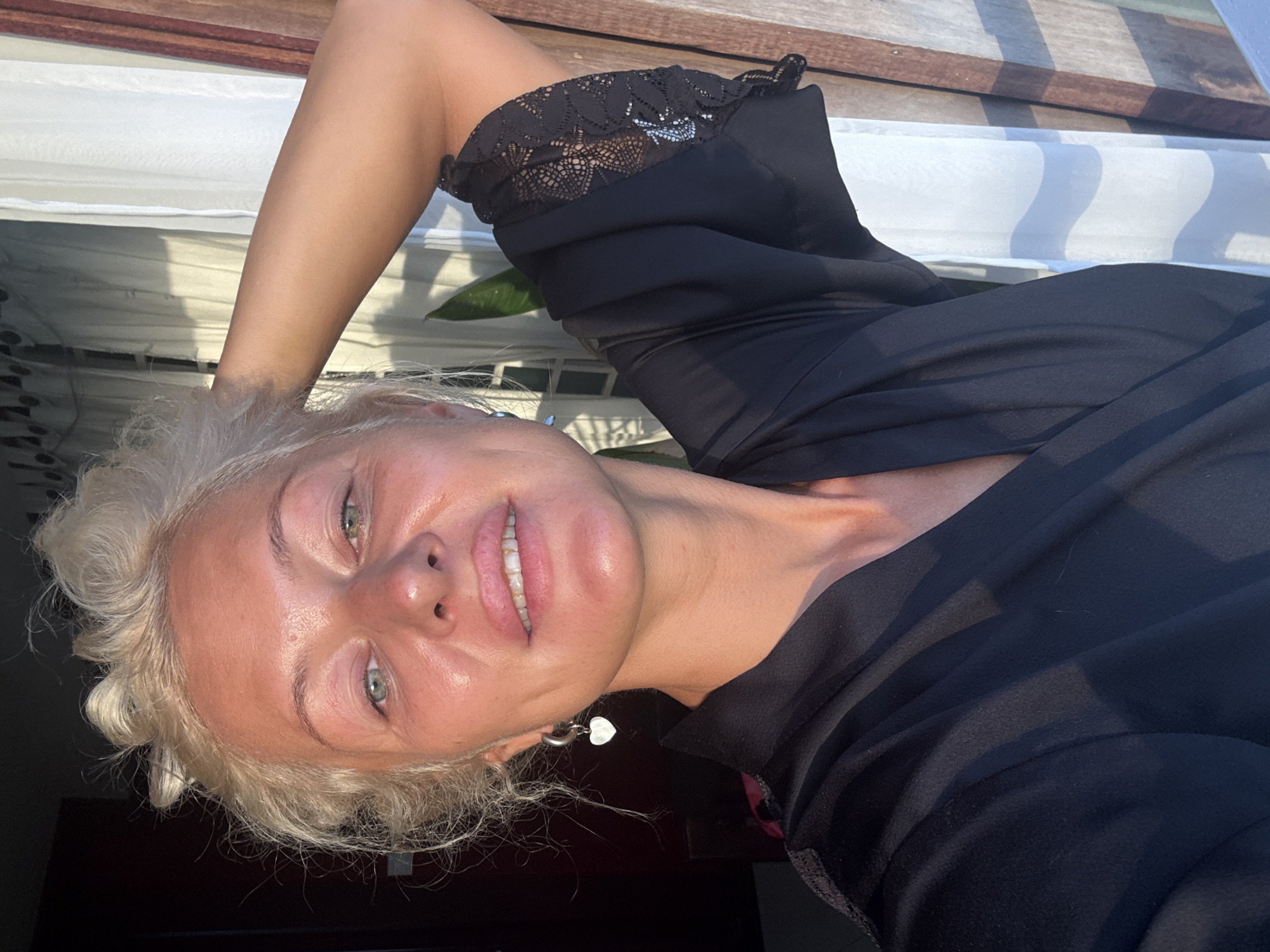 This is a portrait of a blonde woman with fair skin and light blue eyes. She is wearing a black top with lace detailing on the shoulder. The image appears to be a selfie, with the woman's face tilted upwards and her arm extended. Her blonde hair is curly and appears somewhat dishevelled. She has a slight smile on her face and is wearing a heart-shaped earring. The background is blurred, but suggests an outdoor setting with wooden planks and possibly some fabric with a striped pattern. Sunlight is casting shadows and highlighting the contours of her face, indicating it is daytime.