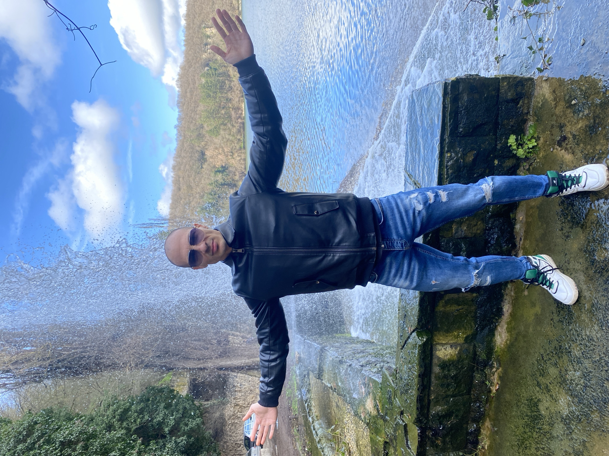 An adult male, bald and wearing dark sunglasses, stands centrally in a park setting within Pleven, Bulgaria, identified as the Pleven Water Cascade in Kaylaka Park. He is positioned on a wet, moss-covered stone pathway, facing the camera with arms extended horizontally and palms open. He wears a black zippered leather jacket, distressed blue jeans with rips at the knees, and white high-top sneakers with green accents. Directly behind him, a multi-tiered artificial waterfall actively cascades water over dark, moss-laden stone structures, generating a dense spray. To the left of the cascade, a rock face is densely covered in green foliage, with a dirt path and partially visible parked vehicles behind it. To the right, a large body of water, likely a lake or pond, extends into the background, reflecting a partly cloudy blue sky. A forested hillside with predominantly bare deciduous trees occupies the distant horizon.