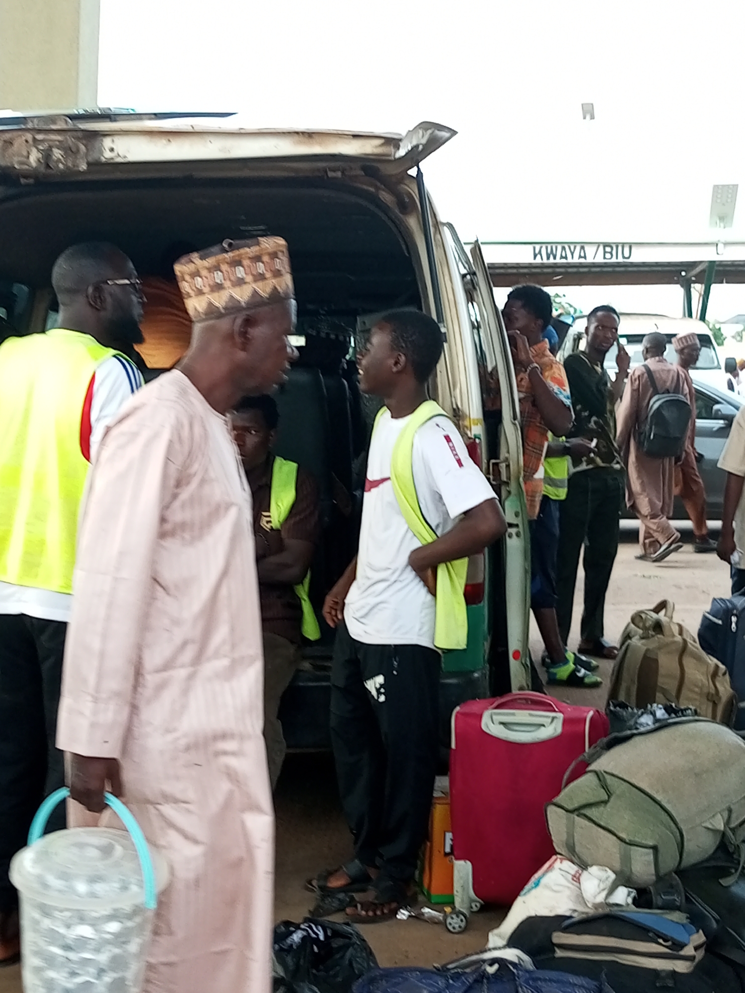 The image depicts a scene at a bus station in Gombe, Nigeria, likely a motor park. Several people, predominantly men, are gathered around a light-colored van with its rear doors open. Many are wearing high-visibility vests, suggesting they might be drivers, conductors, or porters. Luggage, including suitcases, duffel bags, and a bucket, is piled on the ground and near the van. Some individuals appear to be waiting, while others are engaged in loading or unloading the vehicle. The environment suggests an outdoor transportation hub, with a sign in the background indicating "KWAYA / BIU." The overall impression is one of travel and activity, characteristic of a busy motor park in Gombe.
