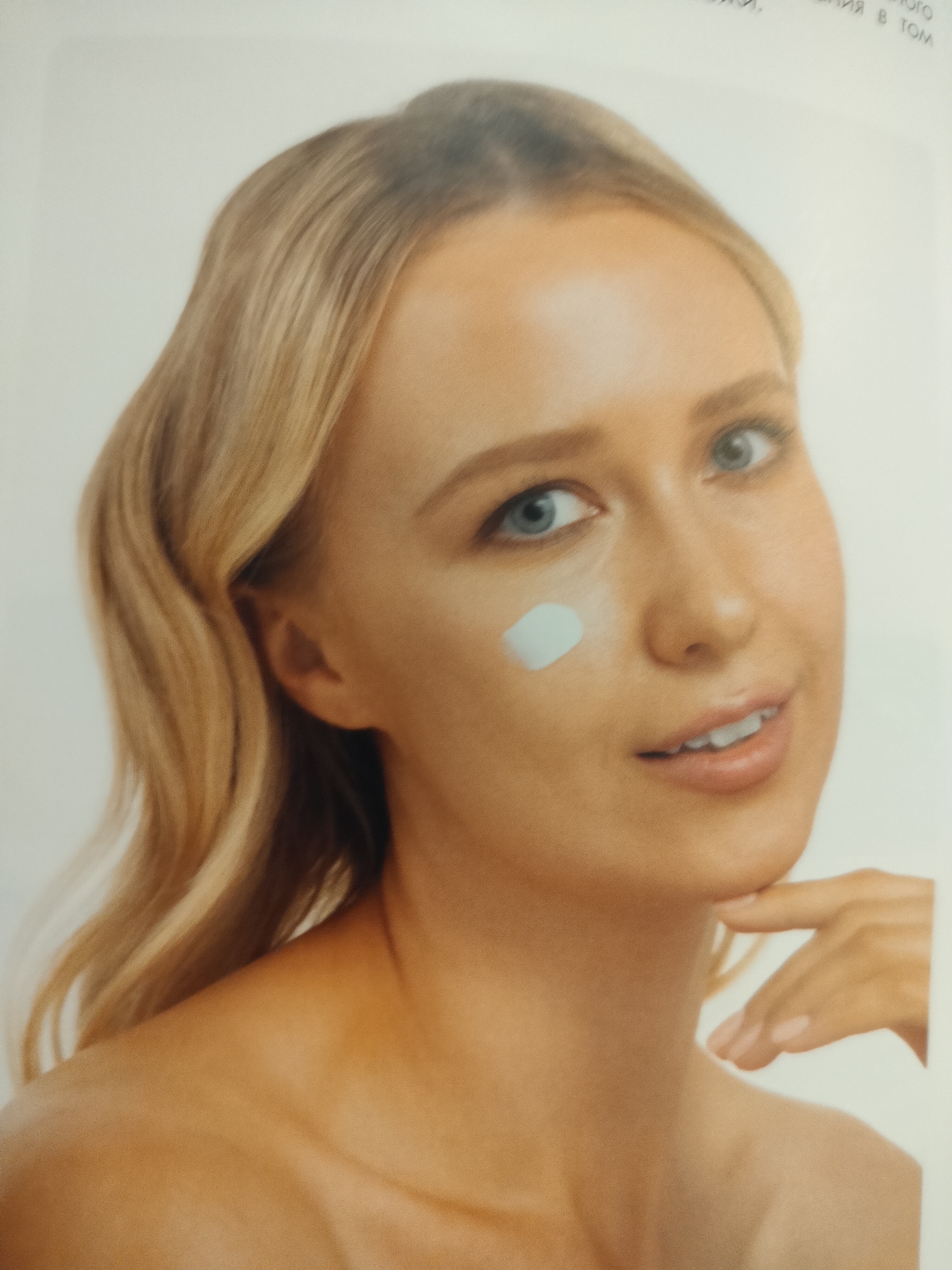 A young Caucasian woman with blonde hair is shown from the chest up against a plain white background. She is looking towards the viewer, her head tilted slightly to her left. A dollop of light blue cream is visible on her left cheekbone. Her right hand is gently touching her chin. The lighting is soft and even, with no discernible shadows. The image does not contain any information or context to suggest a location in the city of Novosibirsk, Russia.