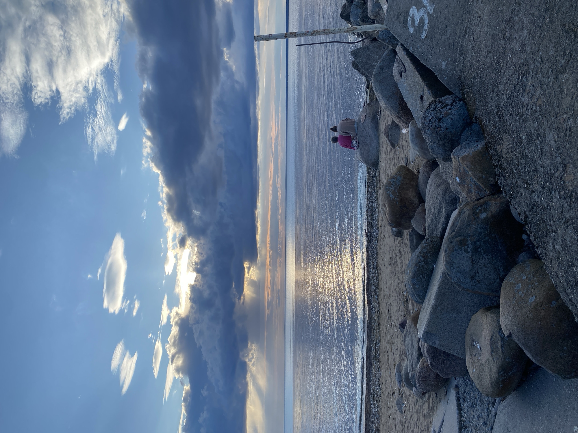 Two individuals are seated on a large boulder overlooking the Gulf of Finland near the Sestroretsk Kurort in Sestroretsk, Russia. The sky is dramatic, with dark, heavy clouds partially obscuring a sun breaking through to cast rays onto the water. The sea is calm, with the sunlight creating a shimmering reflection on its surface. The foreground is composed of a rocky shoreline with large boulders and some sand. A concrete structure with the number "3" painted on it is visible on the right side of the frame, suggesting a man-made element to the shoreline, possibly part of the Sestroretsk Kurort's infrastructure. A weathered wooden post with some attachments stands nearby. The scene evokes a sense of peaceful observation of nature.