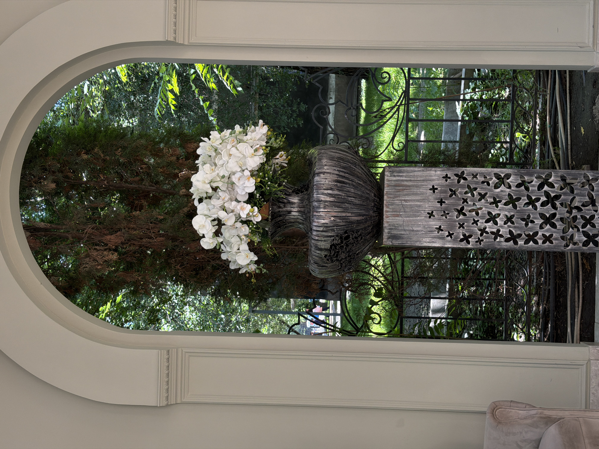 The image, taken in Bishkek City, Kyrgyzstan, shows an indoor arched doorway framing a view of an outdoor garden. In the foreground, a large, textured, grey-toned vase sits on a decorative stand, with white flowers spilling from the vase. Through the archway, a garden with trees, wrought iron fences, and green foliage can be seen. The archway is white and set into a grey wall. A portion of a grey, upholstered chair is visible in the lower right corner.