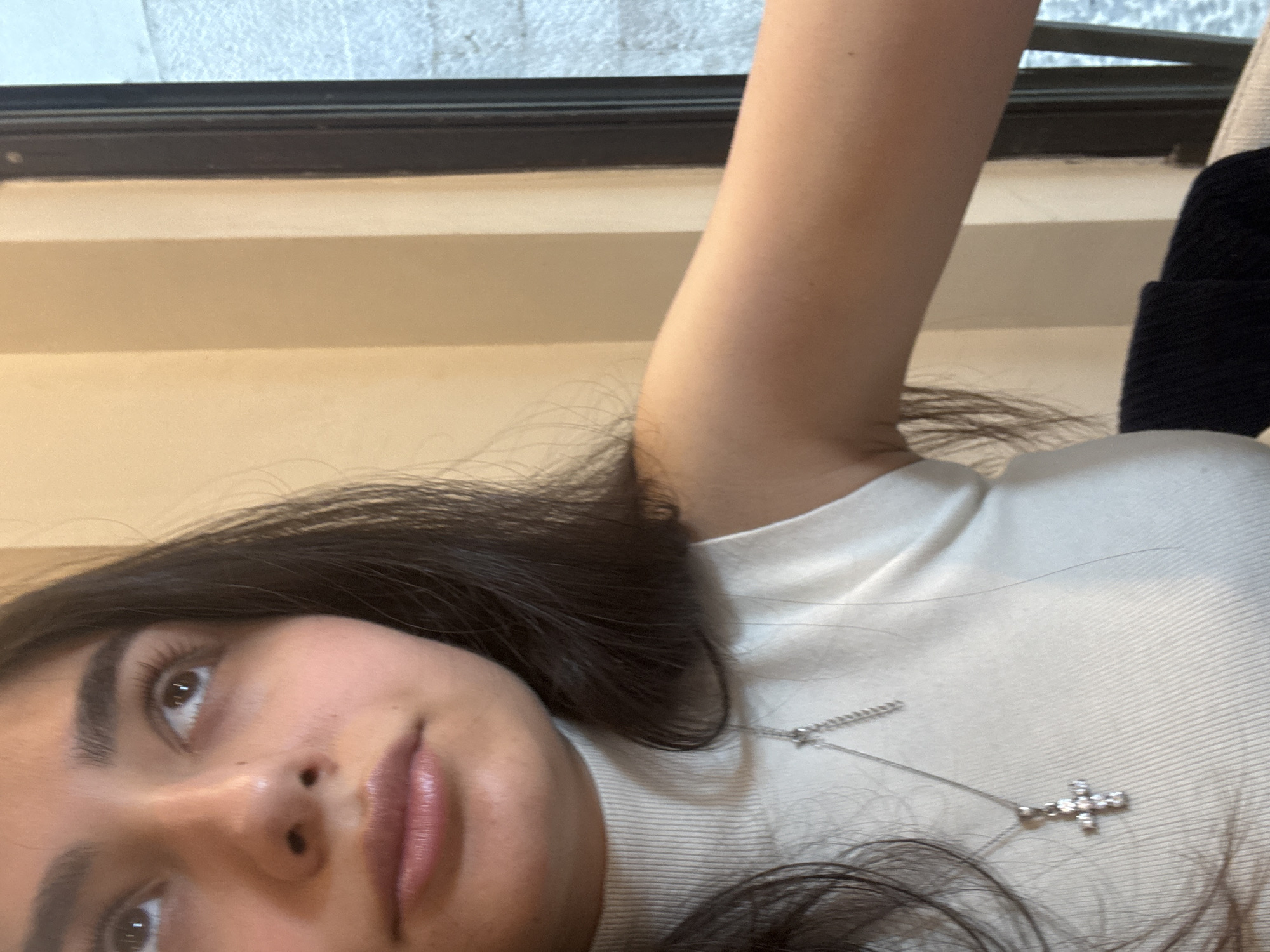 A close-up, portrait-style image, taken from an upward angle, features a young woman with dark brown hair and fair skin. Her head is angled to the left, and she looks upwards with a serene or contemplative expression, her lips slightly parted. She is wearing a light-colored, ribbed turtleneck top. A delicate silver-toned necklace with a small, jewel-studded cross pendant rests on her collarbone area. Her right arm is visible, raised in the upper right portion of the frame. In the background, a dark window frame separates the interior from a blurry view of an exterior building facade, indicating an indoor setting looking out onto an urban environment. The soft, natural lighting suggests it is daytime. No specific activity is depicted beyond posing for the photograph. The scene is situated in Milan, Italy.
