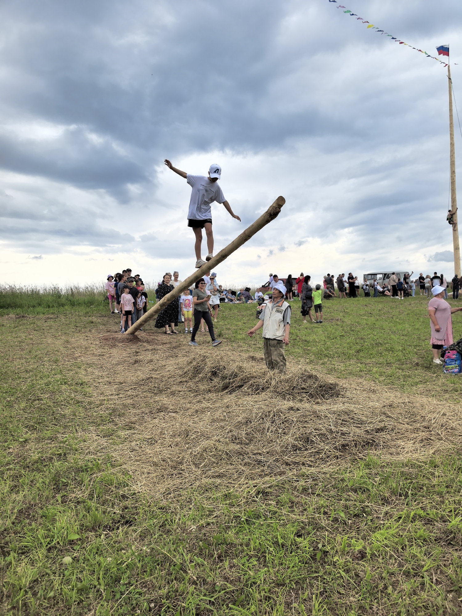In Кояново, Russia, a young person wearing a white baseball cap and a white t-shirt with black shorts is balancing on a slanted log. They are in the center of the frame, with arms outstretched for stability. A crowd of people is gathered on a grassy field, observing the event. In the background, a tall wooden pole is erected, adorned with colorful triangular flags, and topped with a Russian flag. Several individuals are visible on the pole, with one person climbing. The environment consists of a mowed grassy field with patches of dry grass and hay. The sky is overcast with dramatic clouds.