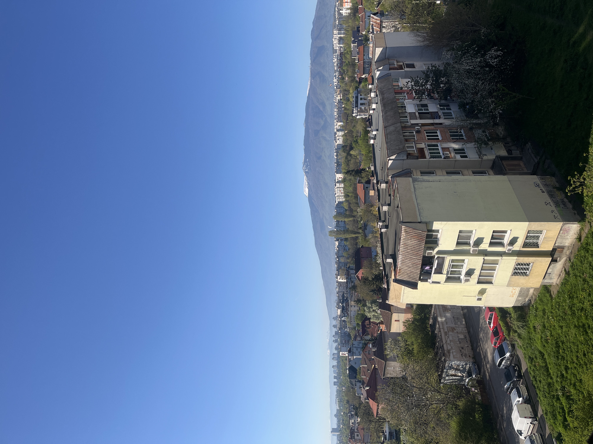 The image shows an elevated view of Sofia, Bulgaria, on a clear, sunny day. The sky is a bright blue. A mountain range is visible in the distance. In the foreground, there are residential buildings of varying heights and colors. Cars are parked along a street next to the buildings. Green grass and trees are also visible.