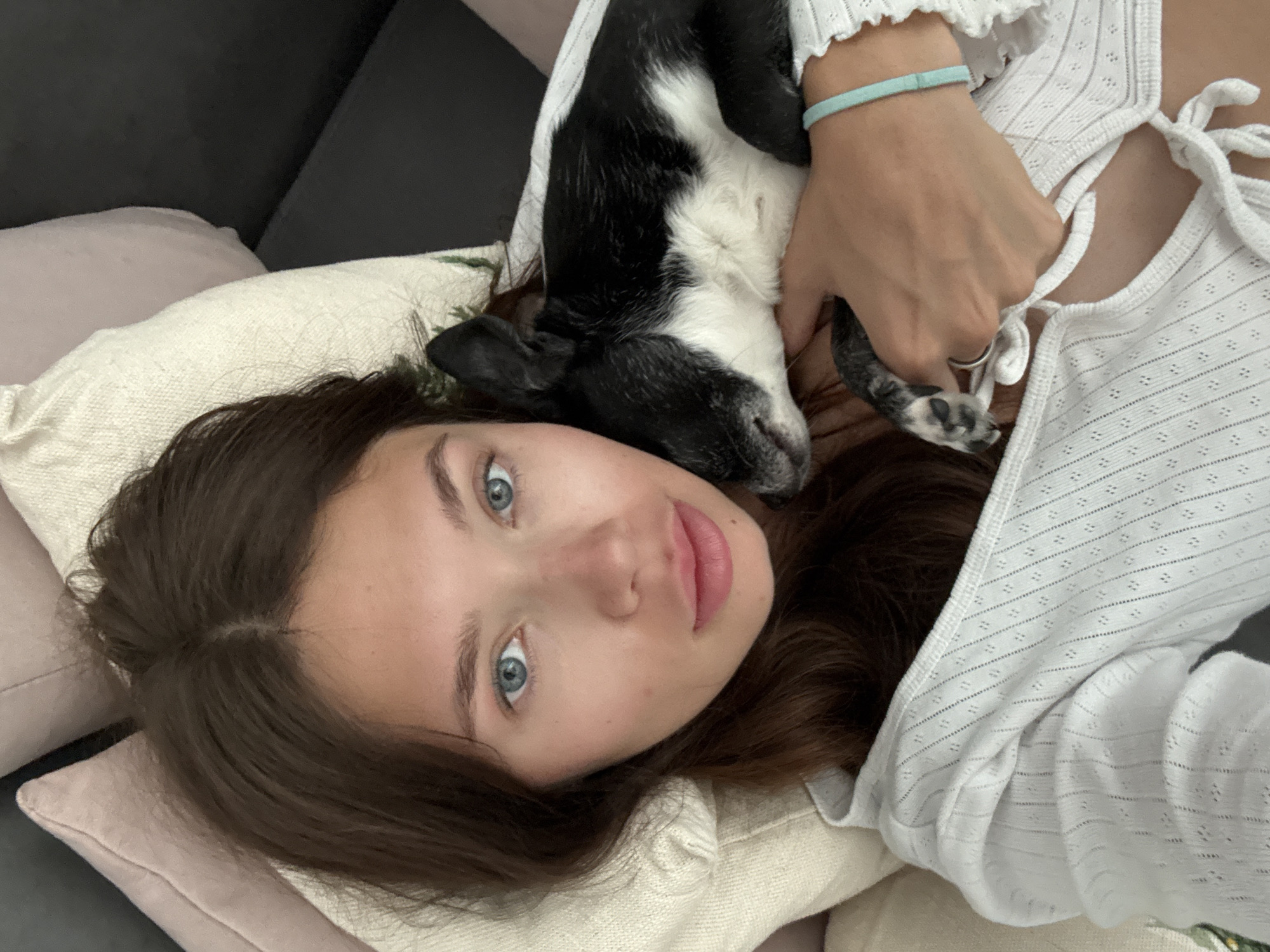 A young woman lies on her back on a light-colored pillow, with a black and white dog resting on her neck and chest. She has brown hair and blue eyes and is wearing a white top. Her right hand is holding the dog. A turquoise bracelet is on her right wrist. The pillow rests on a pink pillow and dark-colored sofa. The location is unknown.