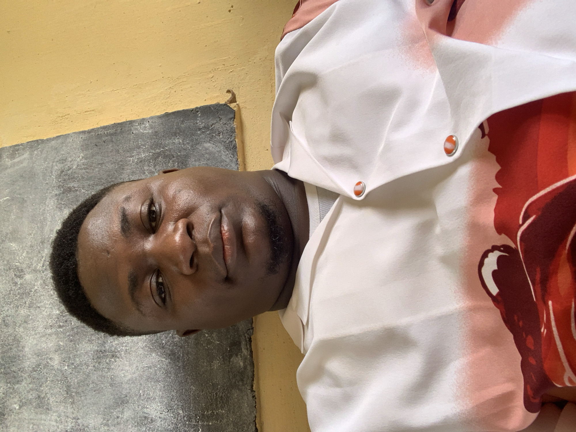 Akwanga, Nigeria: A dark-skinned man with a closely cropped haircut is shown in a medium, close-up shot, facing the camera. He wears a white shirt with orange buttons. The background features a gray, weathered surface and a yellow wall. The lighting appears to be natural.